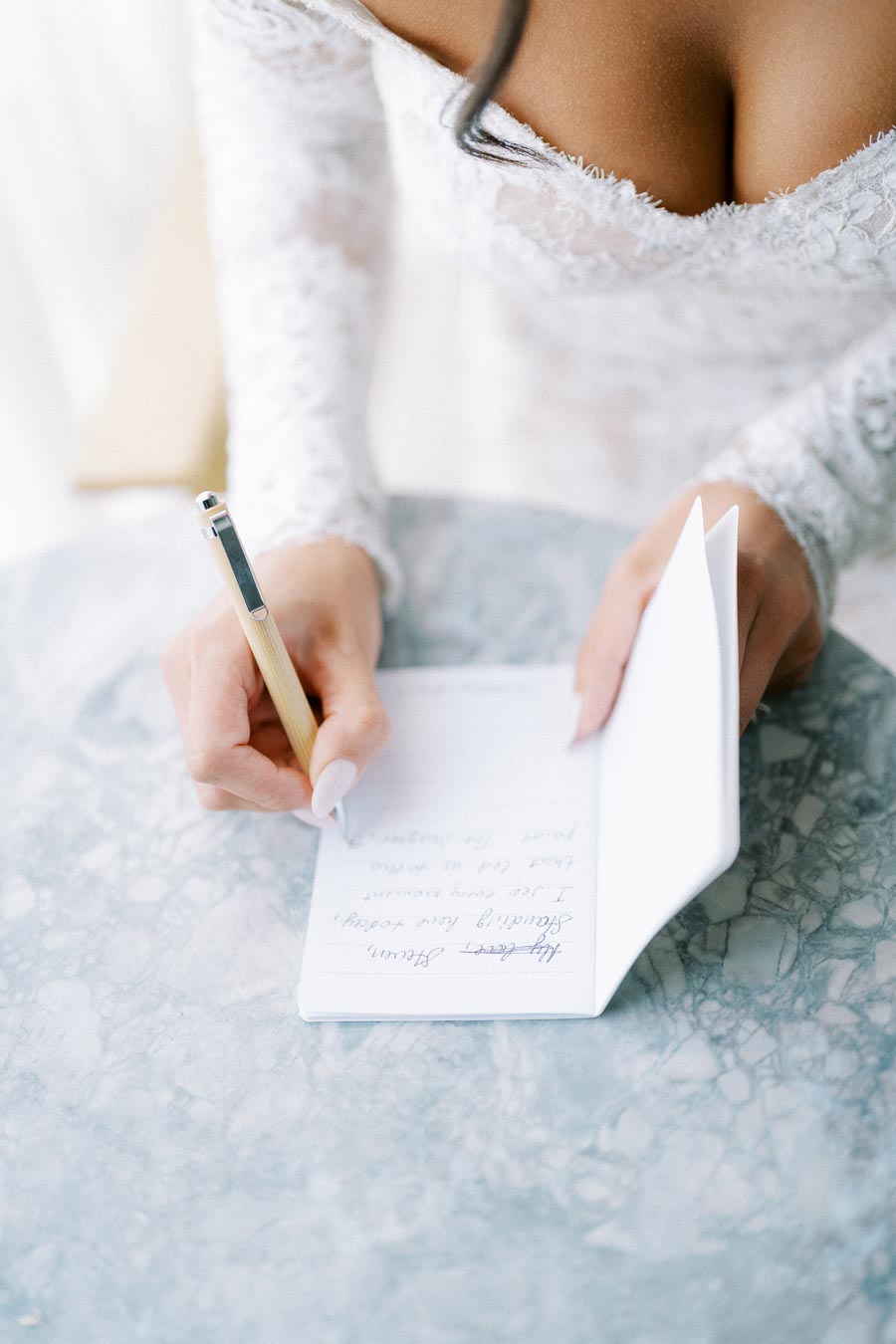 A bride in an elegant lace wedding dress writes heartfelt vows in a notebook on a blue marble table.