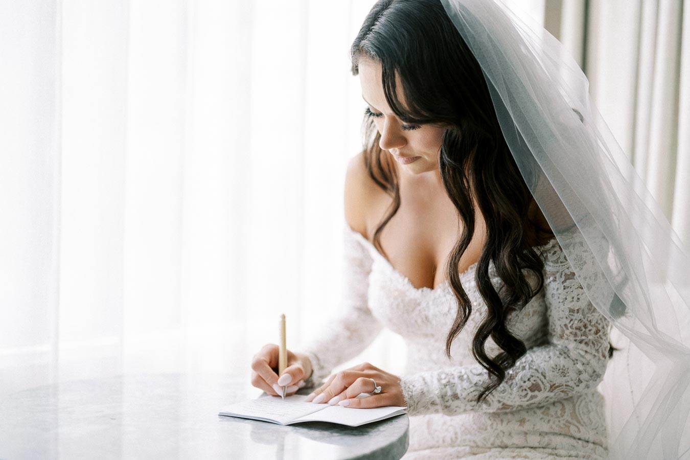 Bride writing in a journal, wearing a lace wedding dress and veil, in a softly lit room with sheer curtains.