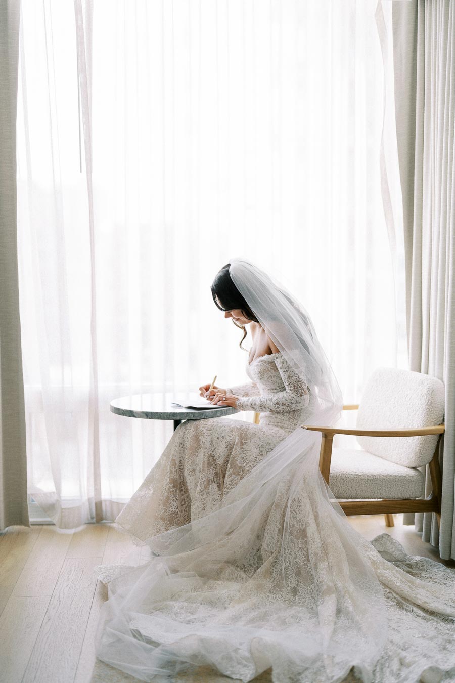 Bride in elegant lace wedding gown writing at a small round table near a window with sheer curtains.