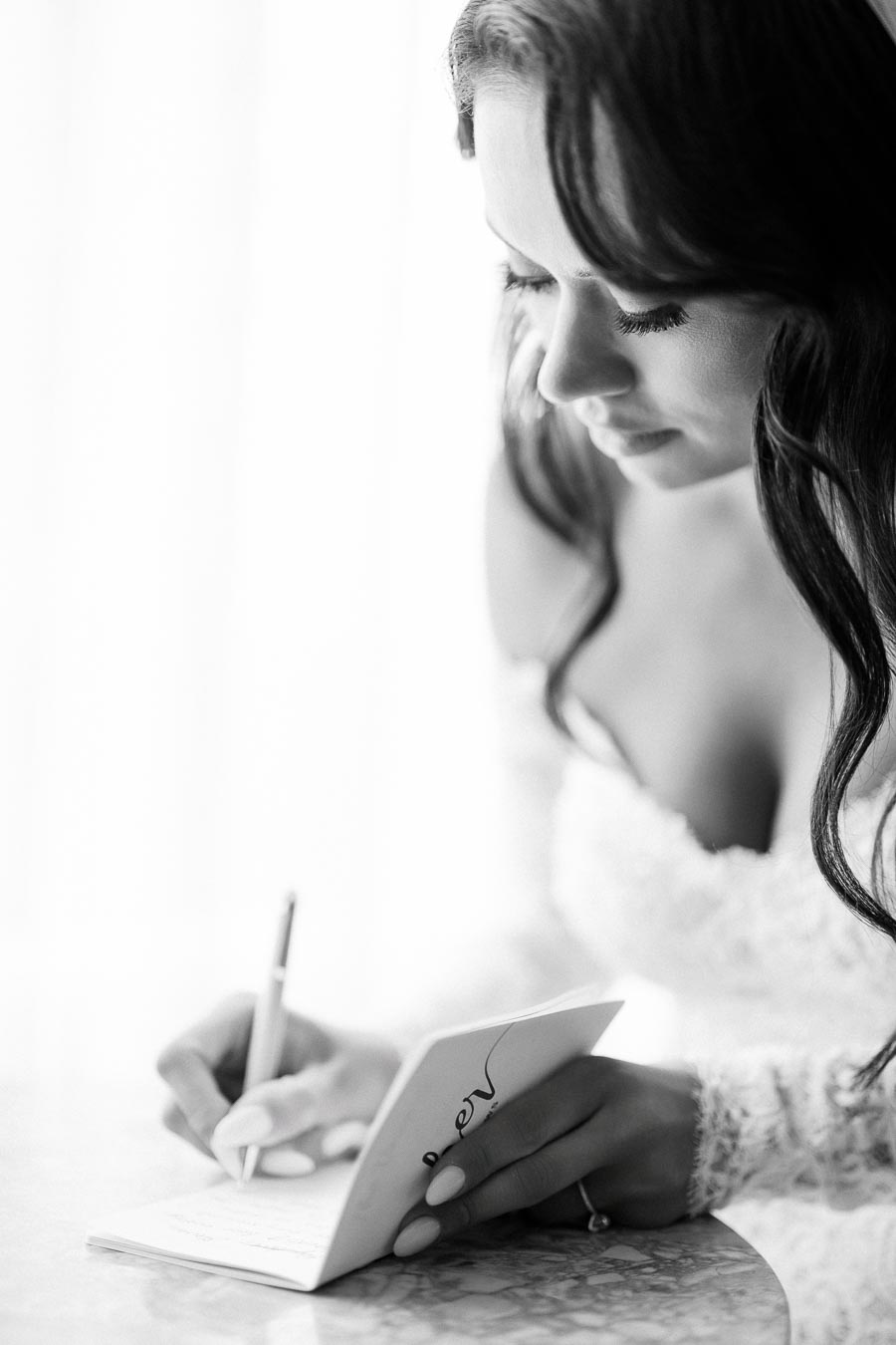 Black and white photo of a bride writing in a notebook at a table, wearing a lace wedding dress and captured in a soft, introspective moment.