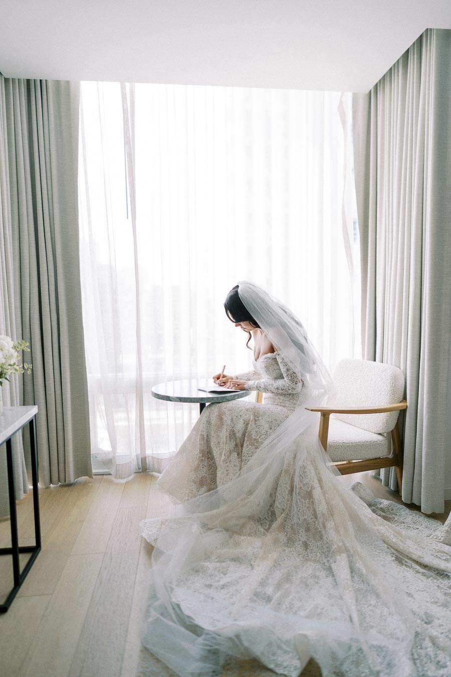 Bride in elegant lace wedding gown sitting by a window, writing a letter before her ceremony, creating a serene and intimate bridal moment.