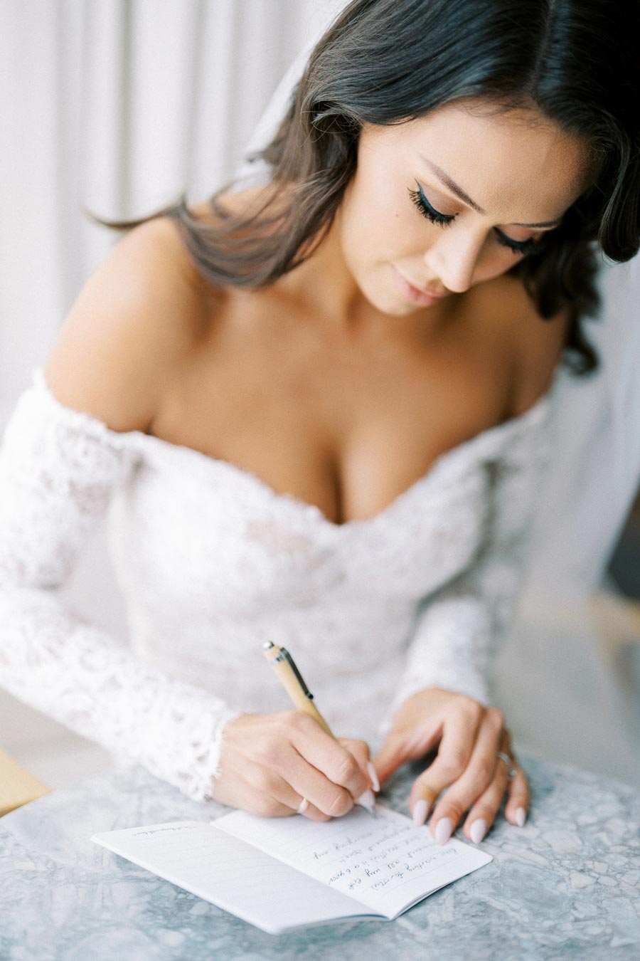 A bride in a white lace wedding dress writing in a notebook at a marble table, capturing a moment of preparation on her wedding day.