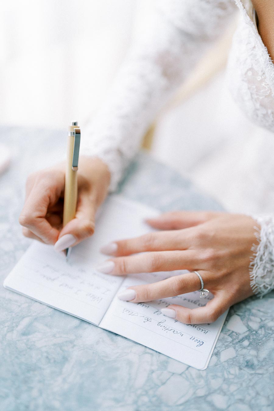 Bride writing in a wedding journal with a pen, showcasing elegant lace sleeves and a diamond ring on a marble table.