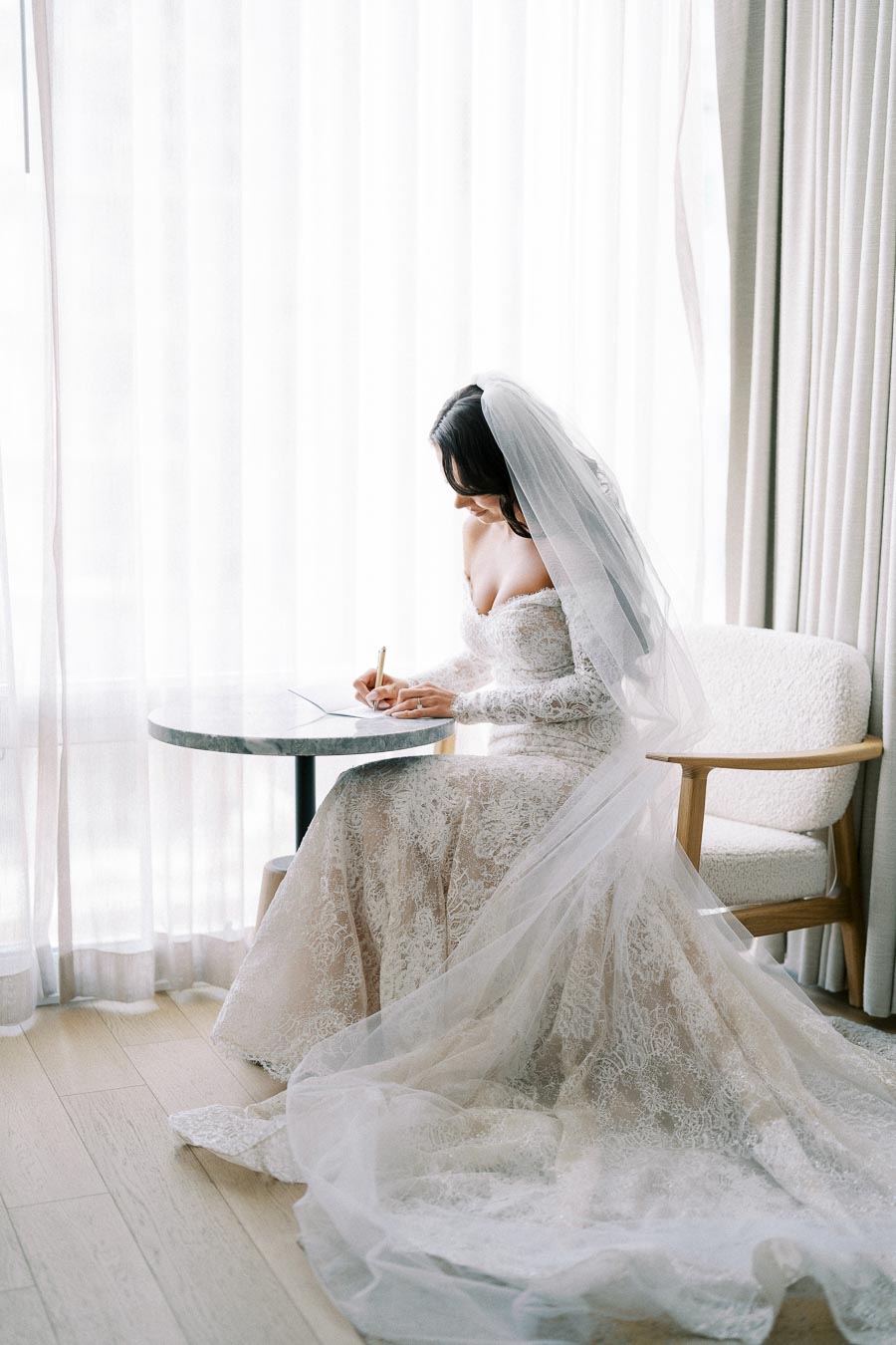 A bride in a lace wedding dress and veil sits by a window with sheer curtains, writing a letter at a small round table, creating a serene and elegant atmosphere.