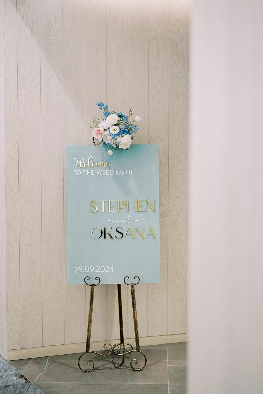 A wedding welcome sign with a bouquet of blue and pink flowers, displayed on an easel, announces the marriage of Stephen and Oksana on September 29, 2024, against a light wood-paneled wall backdrop.