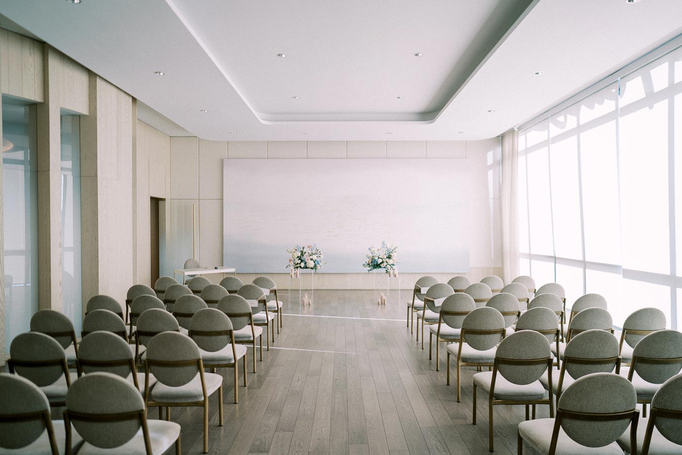 Elegant minimalist wedding ceremony setup in a bright modern venue with rows of chairs and floral arrangements.