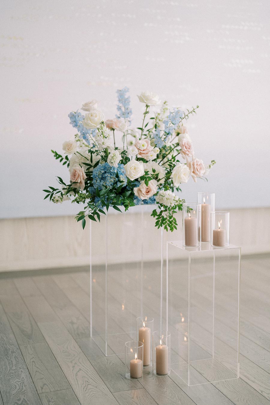 Elegant floral arrangement with pastel blue and pink flowers, accompanied by lit candles in glass holders, placed on clear pedestals against a light background. Ideal for a wedding or romantic event setting.