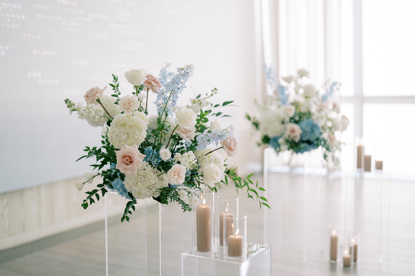 Elegant wedding floral arrangement featuring pastel roses, hydrangeas, and delphiniums accentuated by soft candlelight in a sunlit room, creating a serene and romantic atmosphere.