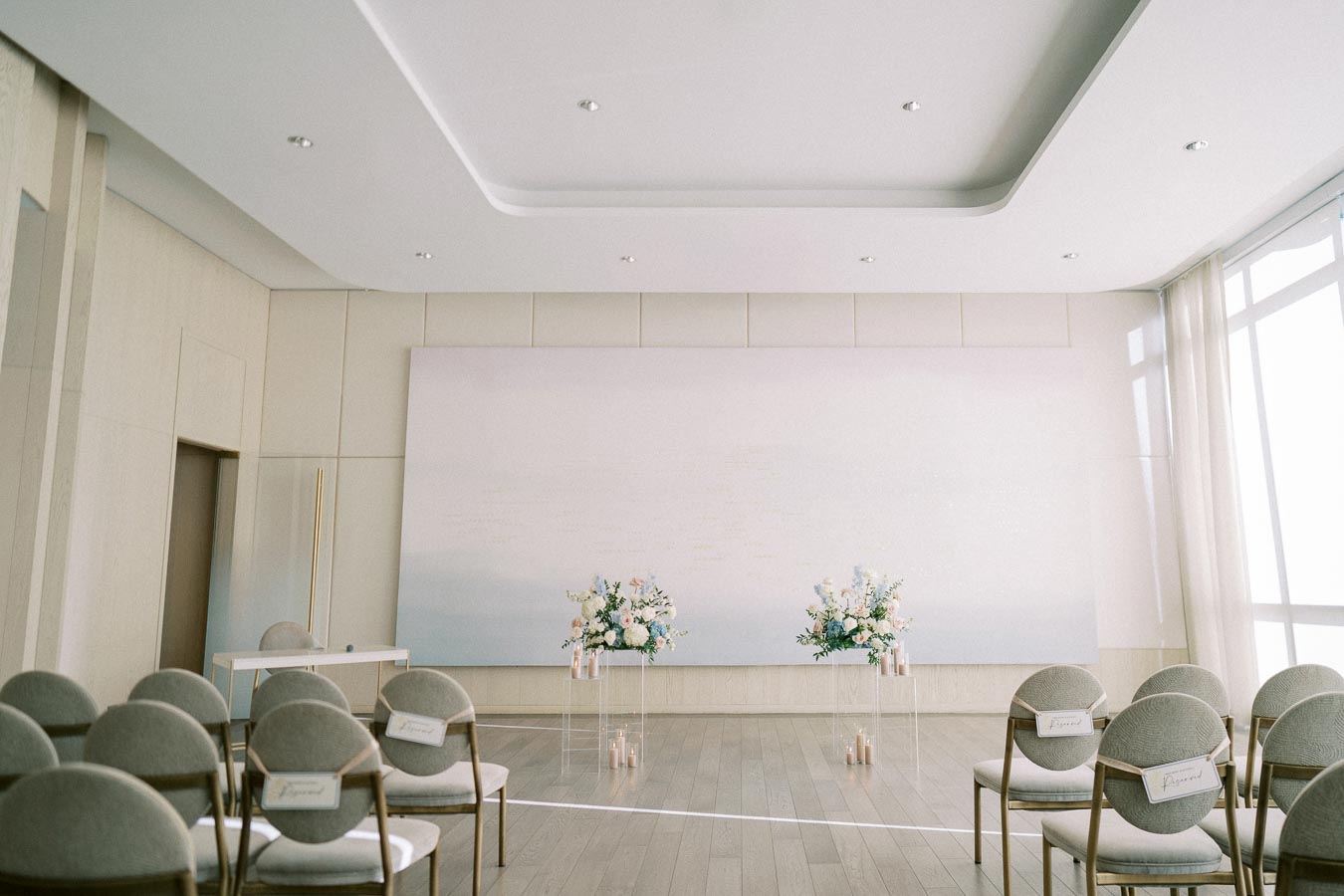 Modern wedding ceremony setup in a bright room with elegant decor, featuring white chairs with reserved signs, floral arrangements, and candles.