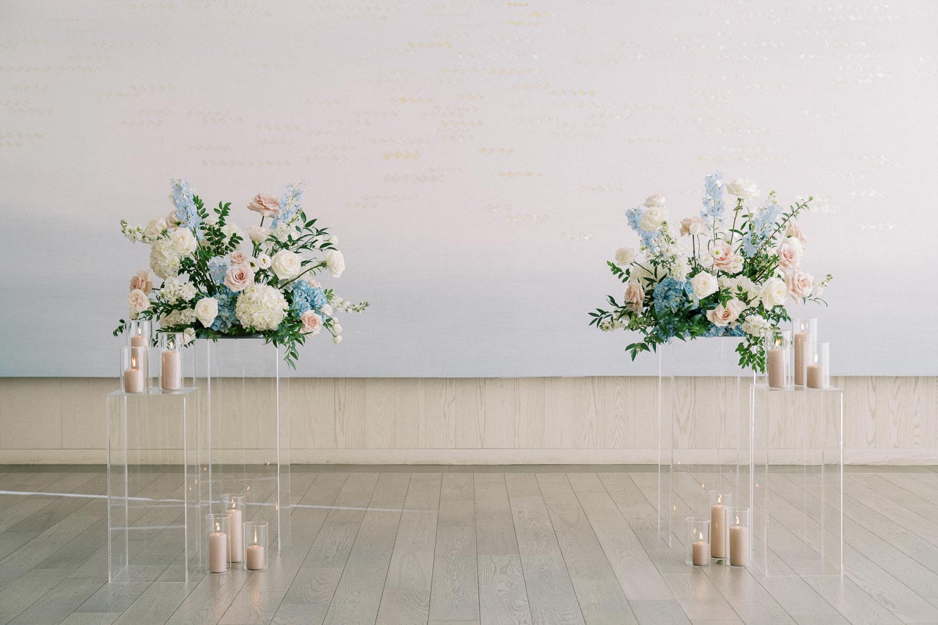 Elegant floral arrangements featuring white and pastel pink roses, blue hydrangeas, and lush greenery on clear stands, accompanied by tall candles in glass holders, set against a soft, neutral backdrop on a wooden floor. Perfect for a wedding or special event decor.