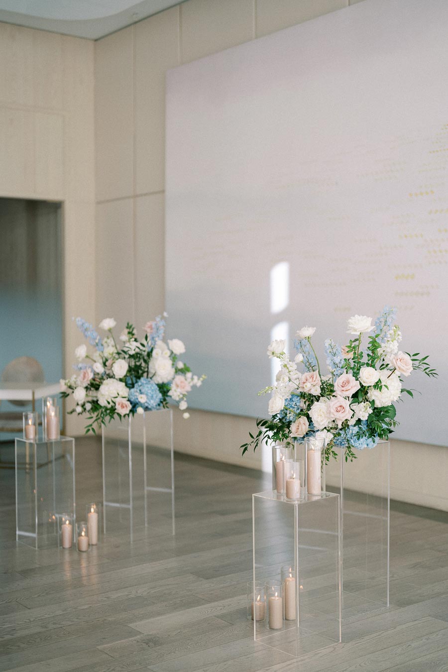 Elegant wedding ceremony setup featuring two floral arrangements with pastel roses and hydrangeas on clear pedestals, surrounded by soft candlelight in a modern venue.