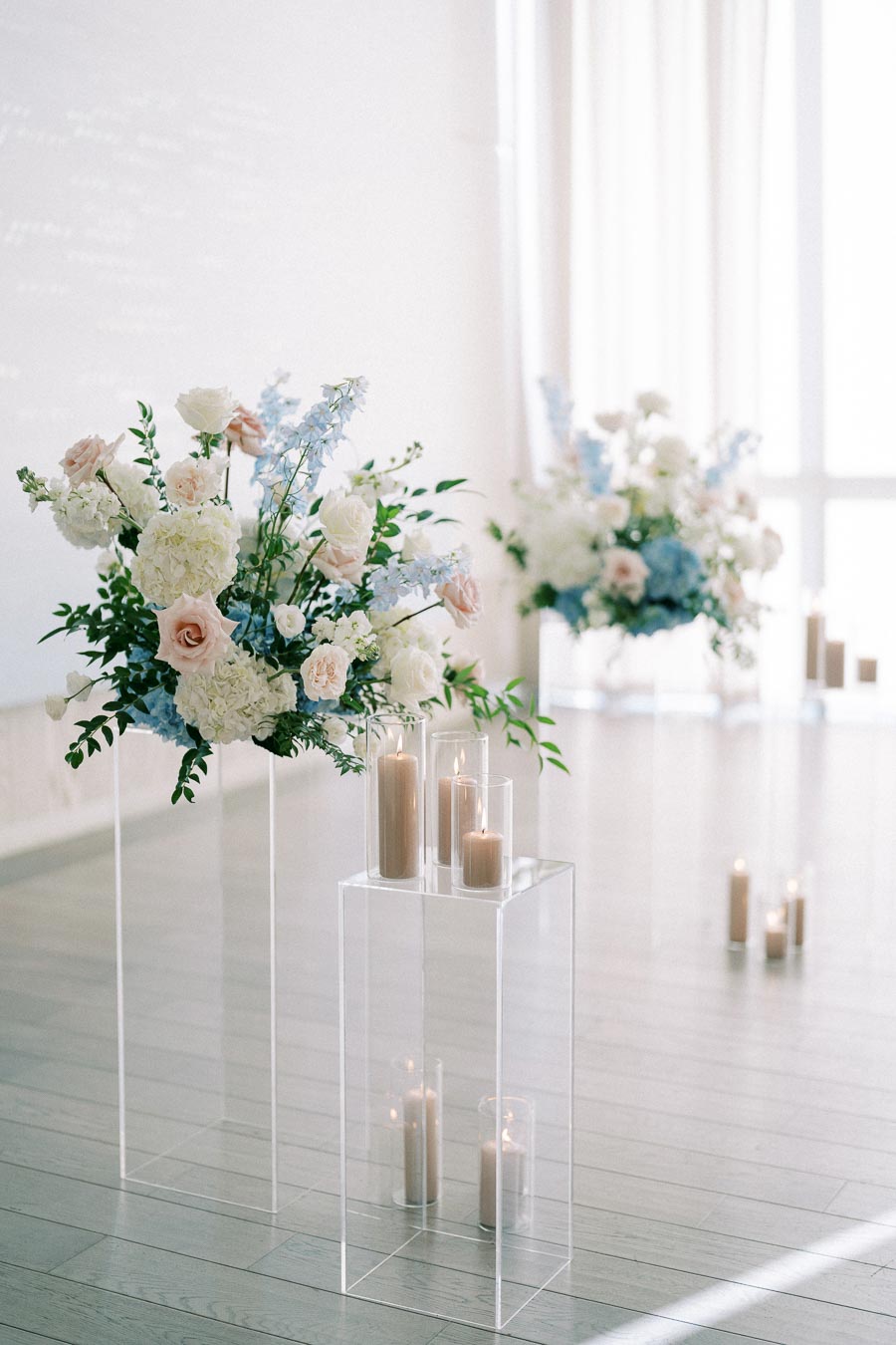 Elegant wedding ceremony setup featuring ivory and pastel floral arrangements on transparent stands, accompanied by lit candles in glass holders, creating a romantic and serene ambiance.