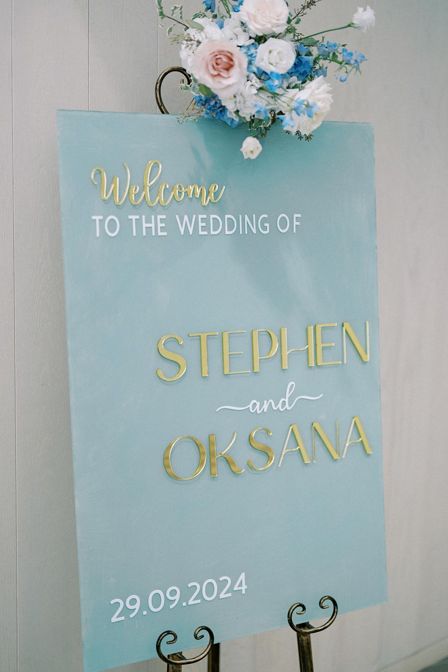 Wedding welcome sign with elegant floral decoration and gold lettering on a teal background, displaying Welcome to the wedding of Stephen and Oksana, 29.09.2024.