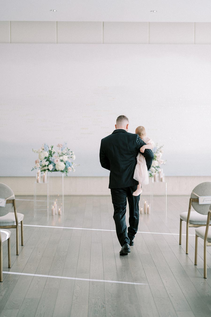 A man in a black suit carrying a young girl down an aisle decorated with flowers and lit candles in a bright, elegant wedding venue.