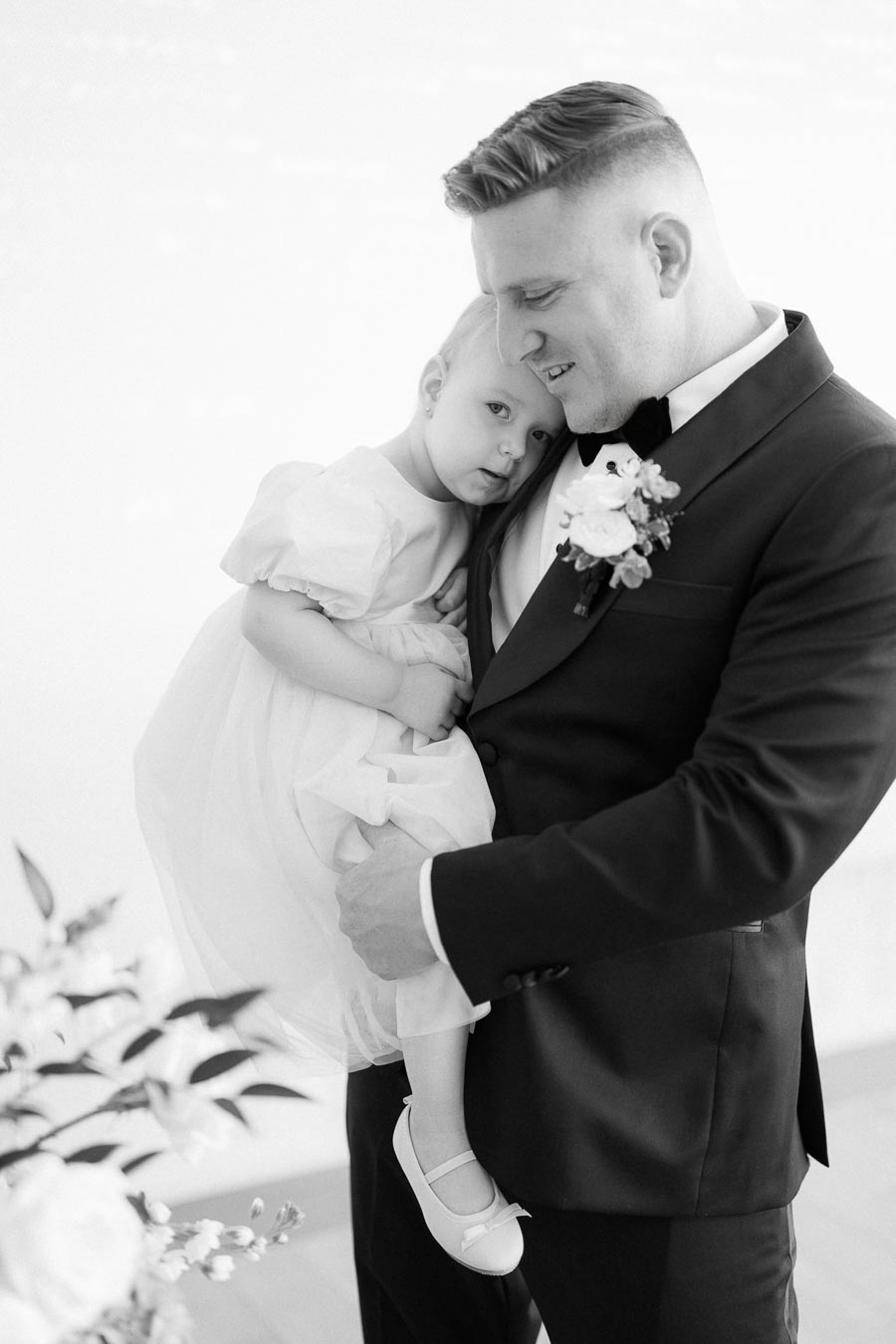 Father holding daughter at formal event, little girl in white dress snuggles close, man in black suit with boutonniere smiles, tender family moment.