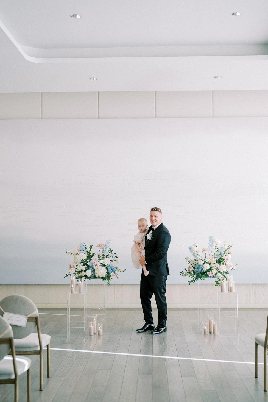 A well-dressed man holding a baby at a modern indoor wedding ceremony with elegant floral arrangements and candles.