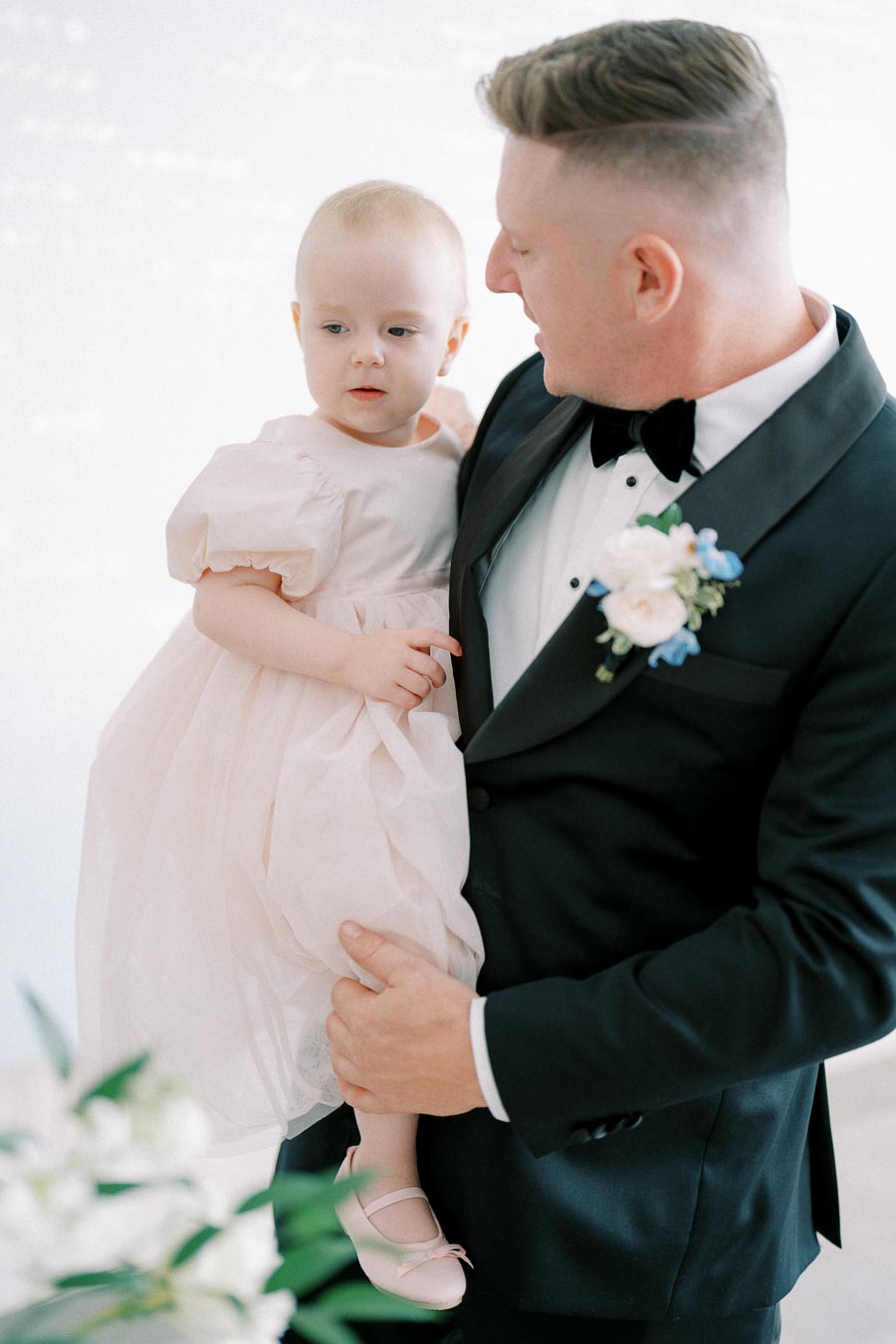 A man in a formal black tuxedo holding a young child dressed in a pink dress during an elegant event with soft lighting.