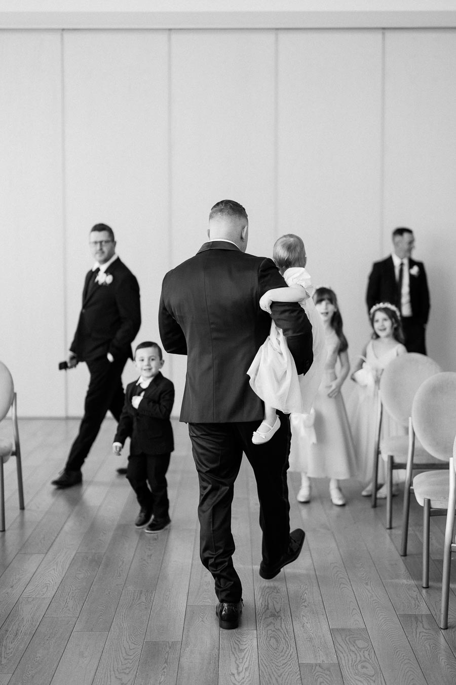 Black and white photo of a man in a suit carrying a child, walking down an aisle with other children and individuals also dressed in formal attire, in an indoor setting.
