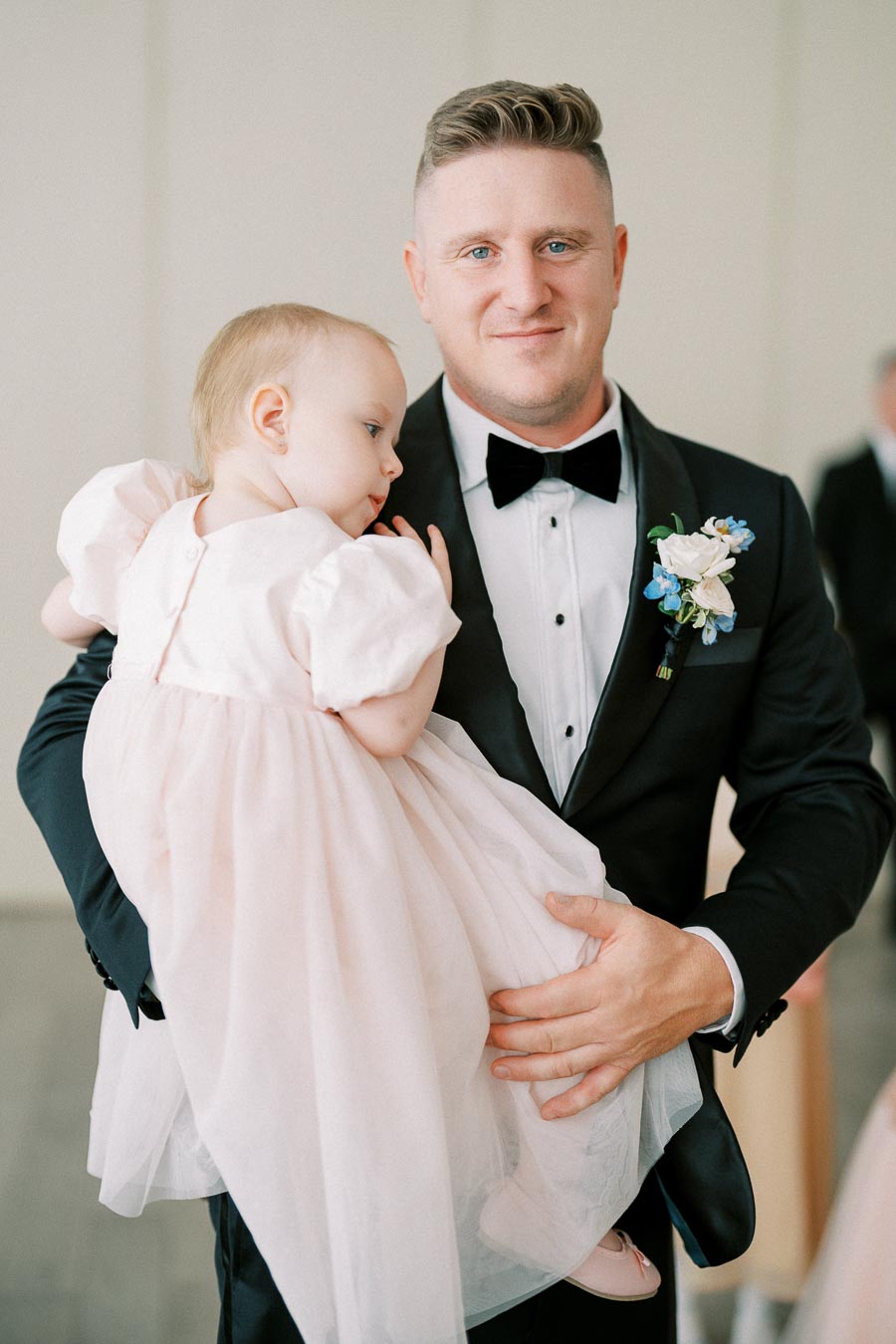 A man in a formal black tuxedo holding a baby girl in a pink dress.