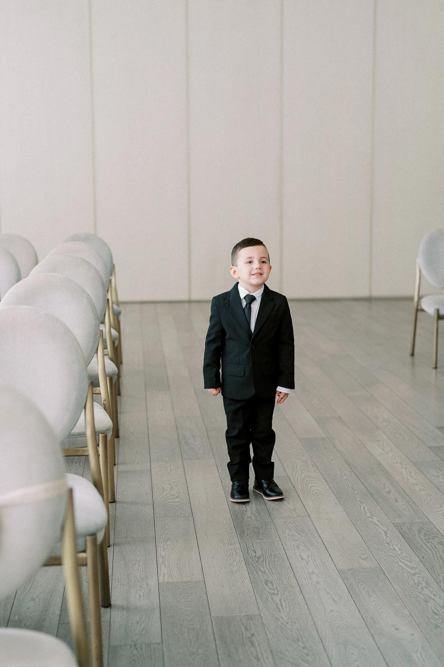 A young boy in a black suit and tie standing in an empty room with rows of chairs.