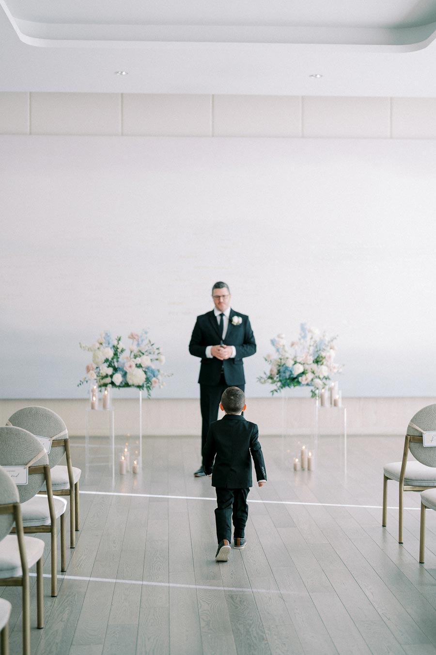 A young boy in a suit walks down a modern, minimalist wedding aisle towards an adult in formal attire, with elegant floral arrangements and lit candles decorating the intimate indoor setting.