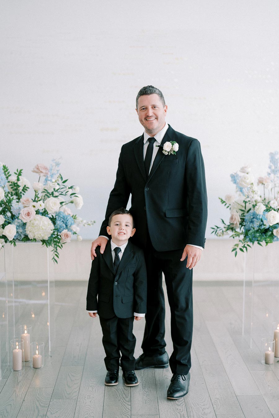Adult and child in matching black suits pose with floral arrangements in an elegant setting; candles adorn the floor, adding warmth to the sophisticated atmosphere.