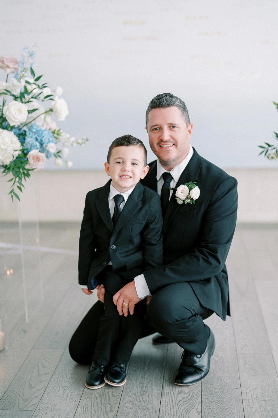 Father and young son in matching suits at a wedding, posing happily next to floral arrangements.