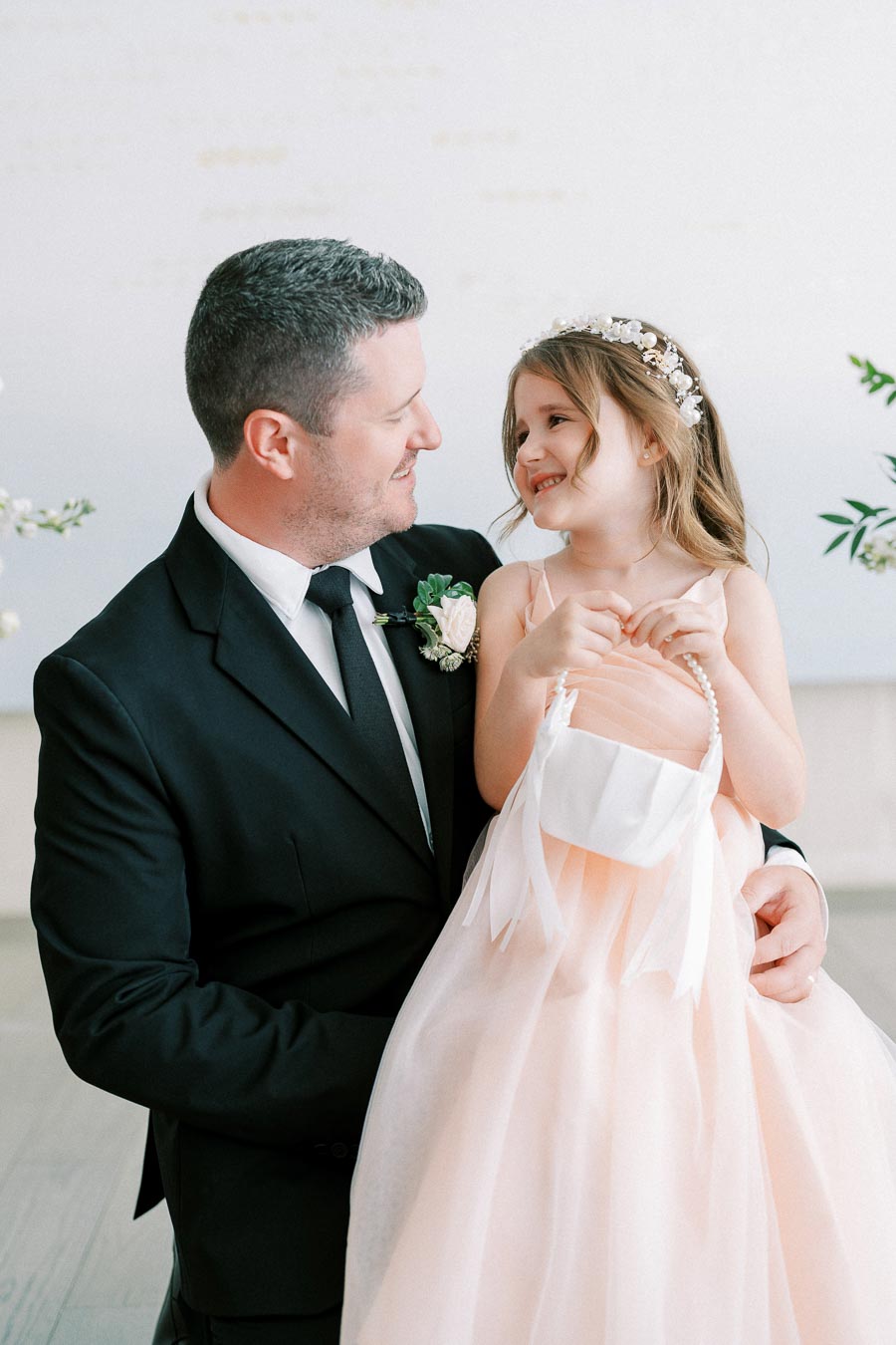 Father in a black suit smiling at his daughter in a peach dress, holding a flower girl basket, during a wedding ceremony.