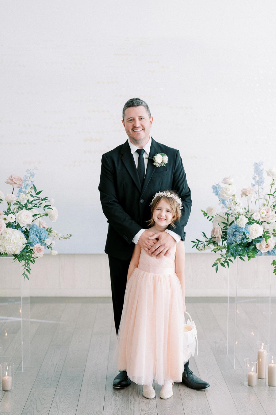 A man in a black suit and tie, adorned with a boutonniere, stands smiling with a young girl in a pale pink dress and floral headband. They are surrounded by elegant floral arrangements of white and blue flowers, with candles on the floor, set against a light background.