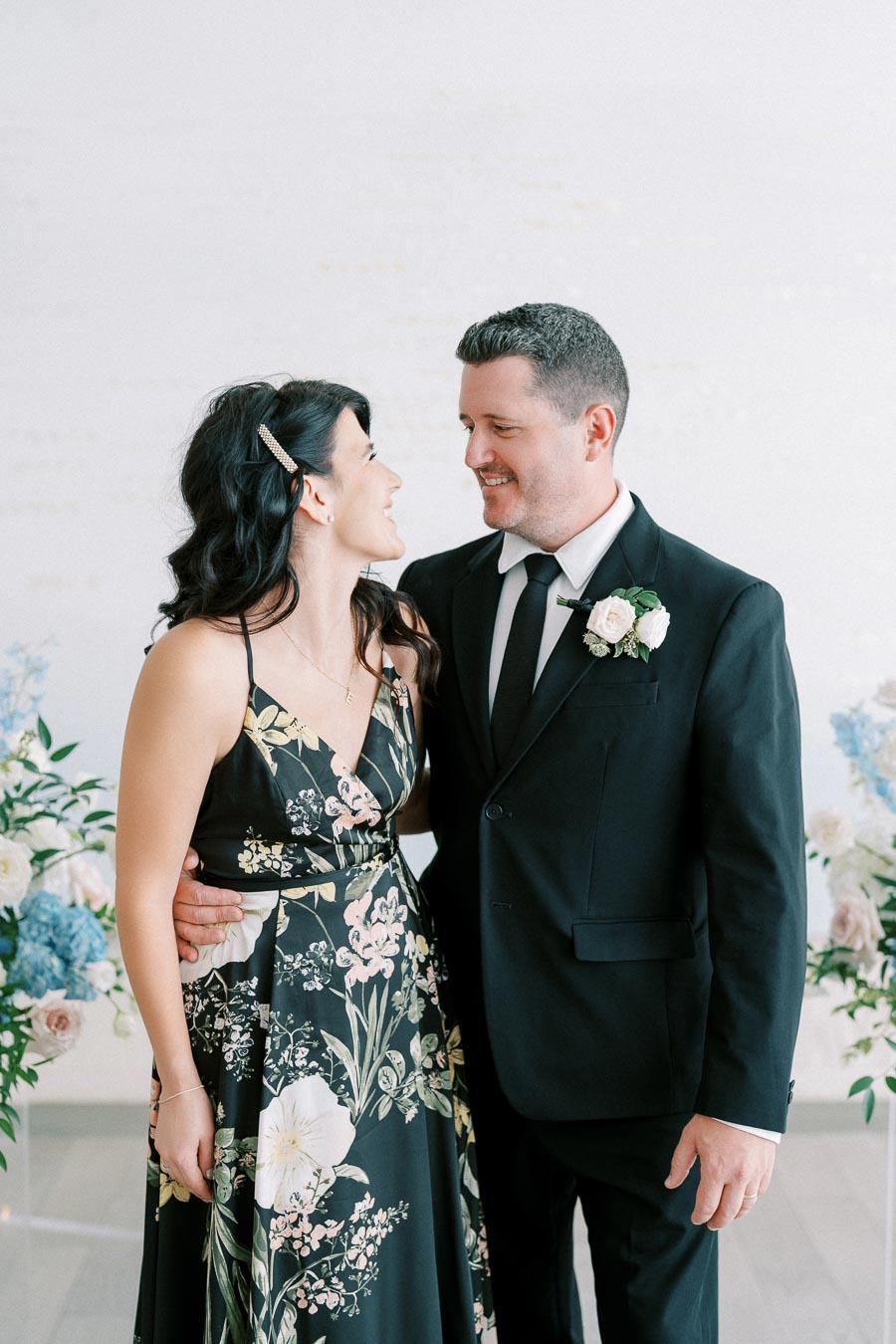 A couple smiling at each other, dressed in formal attire; the woman wears a floral dress and the man is in a black suit with a boutonniere, standing in front of a light background with floral arrangements.