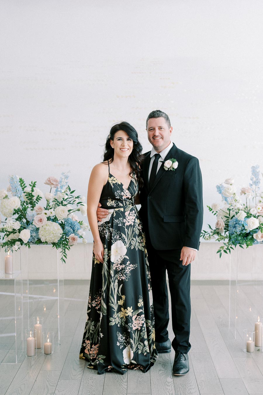 A couple dressed in formal attire stands smiling in a beautifully decorated room with floral arrangements and candles. The woman wears a floral-patterned dress and the man wears a black suit with a boutonniere.