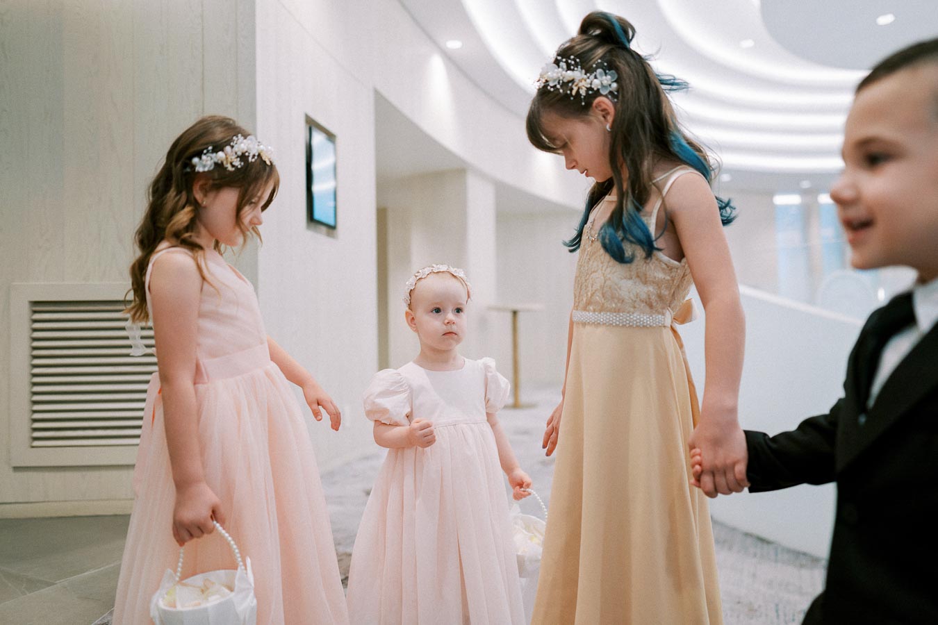 Children dressed elegantly at a wedding, with girls in pastel dresses and floral headbands, and a boy in a suit, standing in a modern venue.