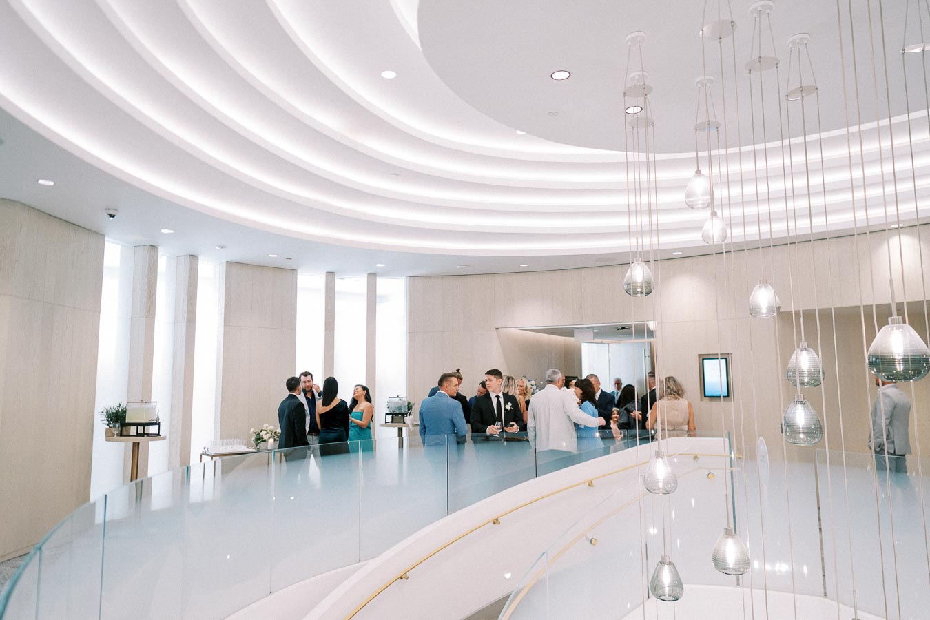 People gathered in a modern, elegantly designed conference room with curved ceiling lights and a glass railing, having conversations during an event or networking session.