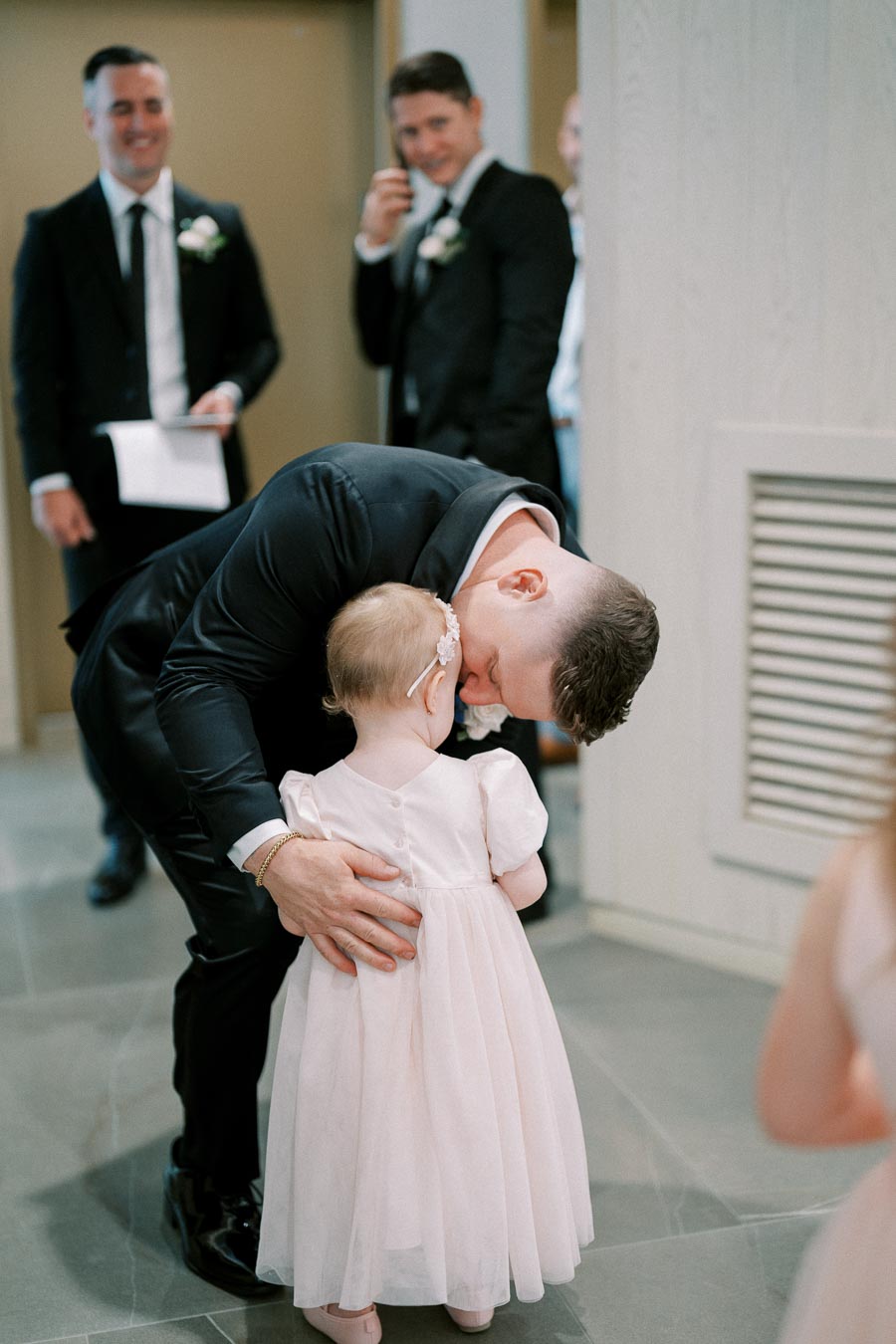 Man in formal suit affectionately embraces a young girl in a pink dress during a wedding ceremony, with smiling guests in the background.