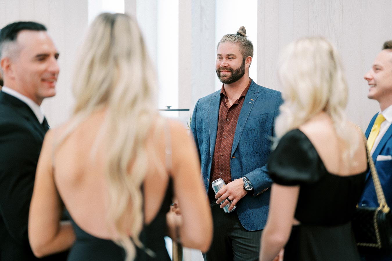 A group of elegantly dressed people engaging in conversation at a formal event, with one person in a blue suit holding a beverage can.