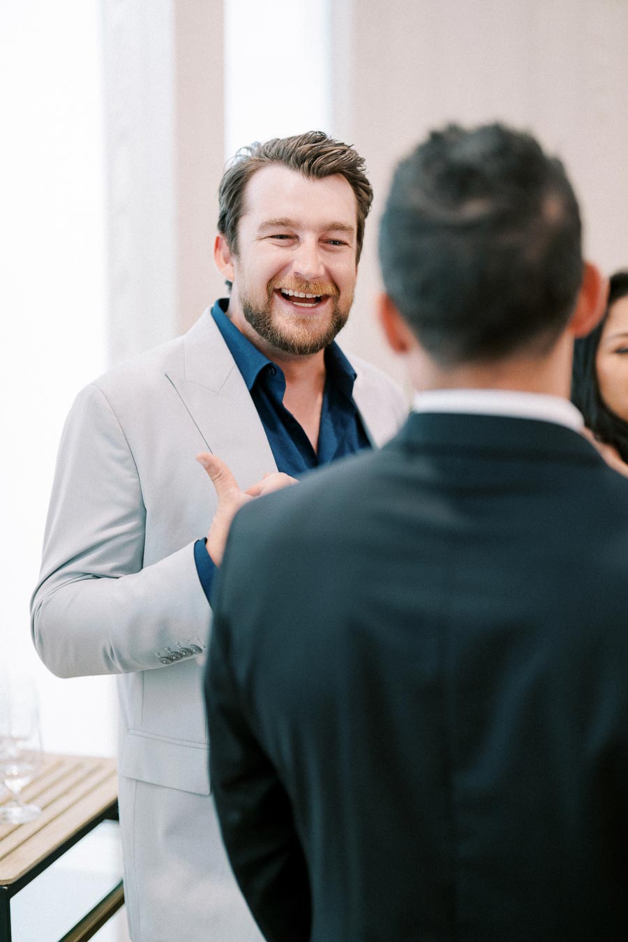 Man in light suit smiling and engaging in conversation at a social gathering with another person in the foreground.