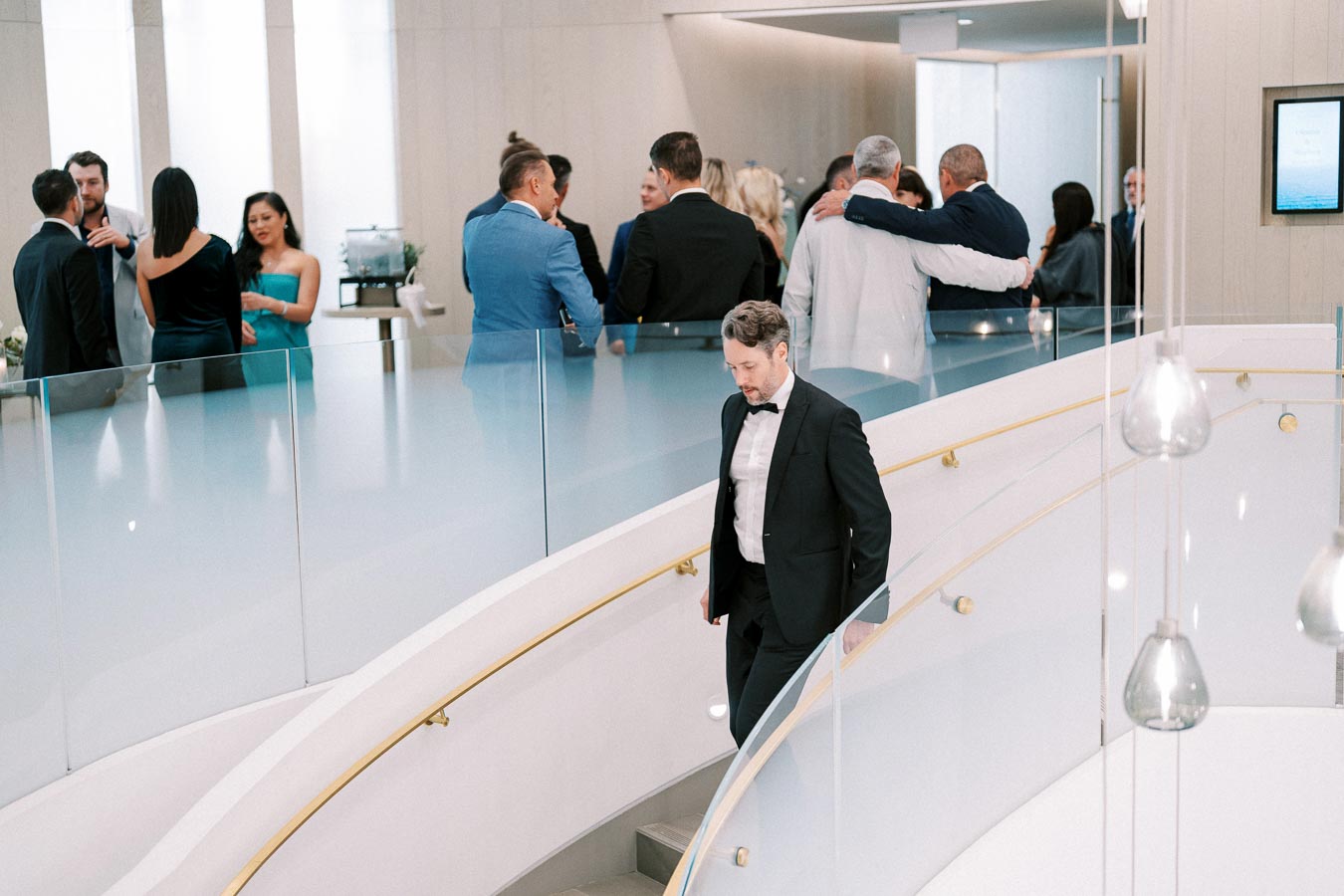 A man in a formal black tuxedo walks down a curved staircase at a stylish indoor event, with a group of elegantly dressed people socializing in the background, creating a sophisticated and upscale atmosphere.