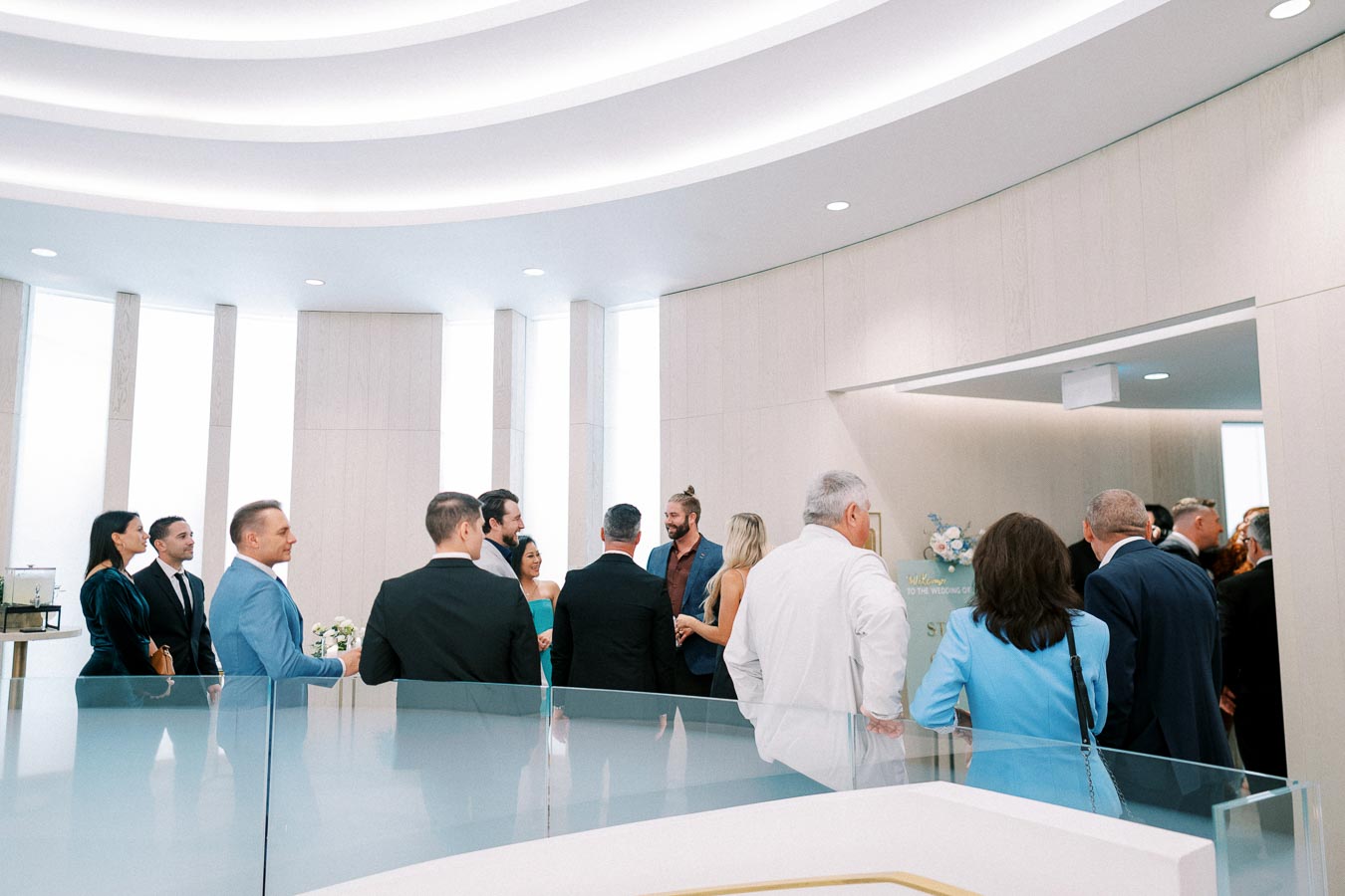 A group of elegantly dressed people gather in a modern, well-lit conference room with sleek design and glass accents, engaging in conversation during a formal event or ceremony.