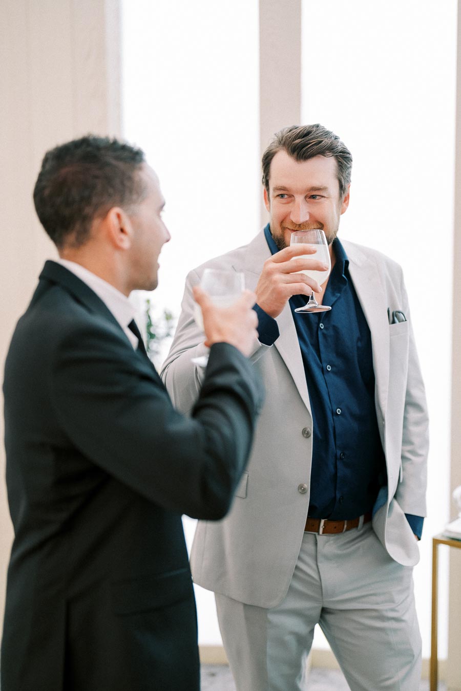 Two men in formal attire enjoying a conversation over drinks, one wearing a light gray suit and the other in a black suit, in a bright indoor setting.