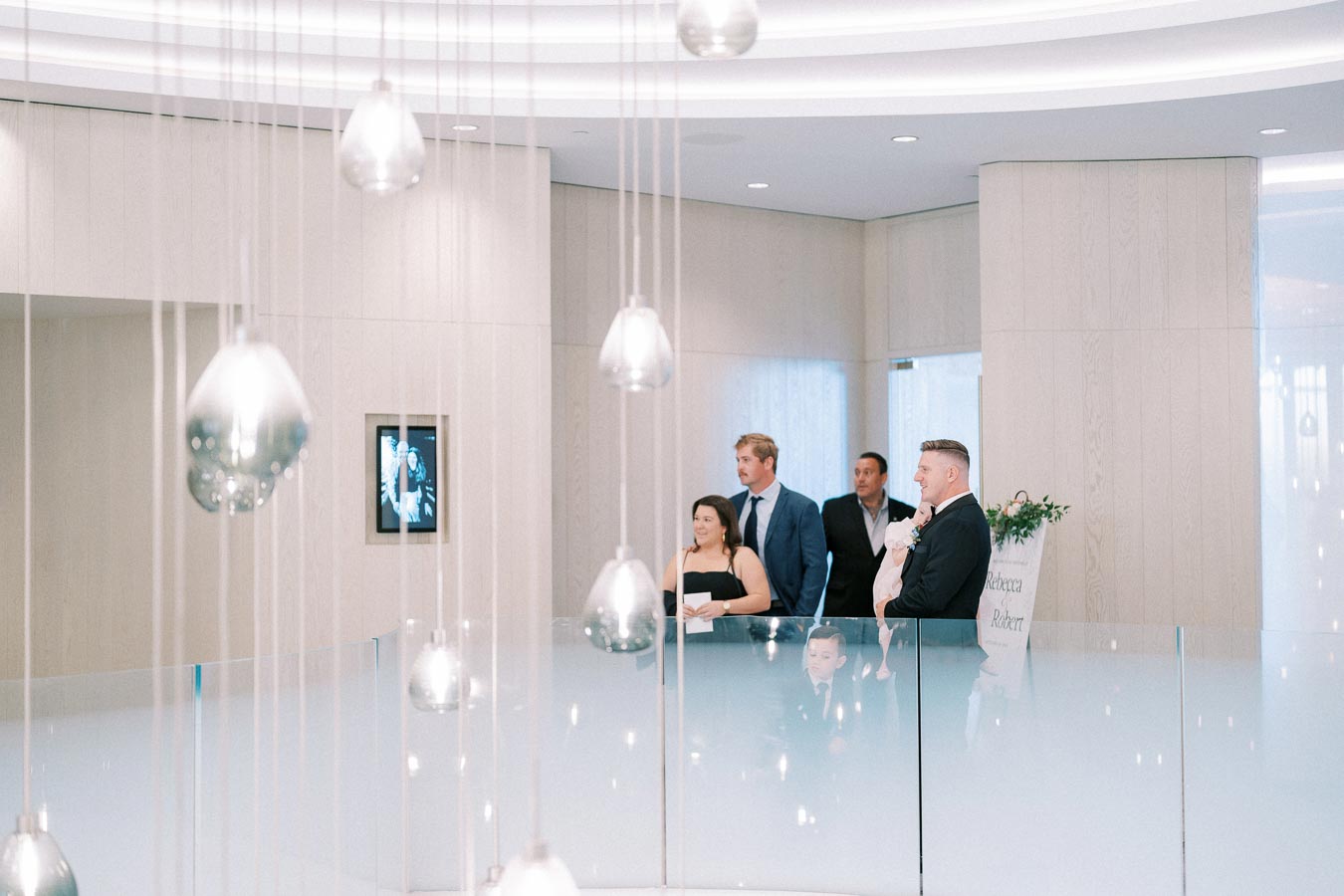 A group of elegantly dressed people standing in a modern, well-lit venue with decorative hanging lights and a framed sign in the background.