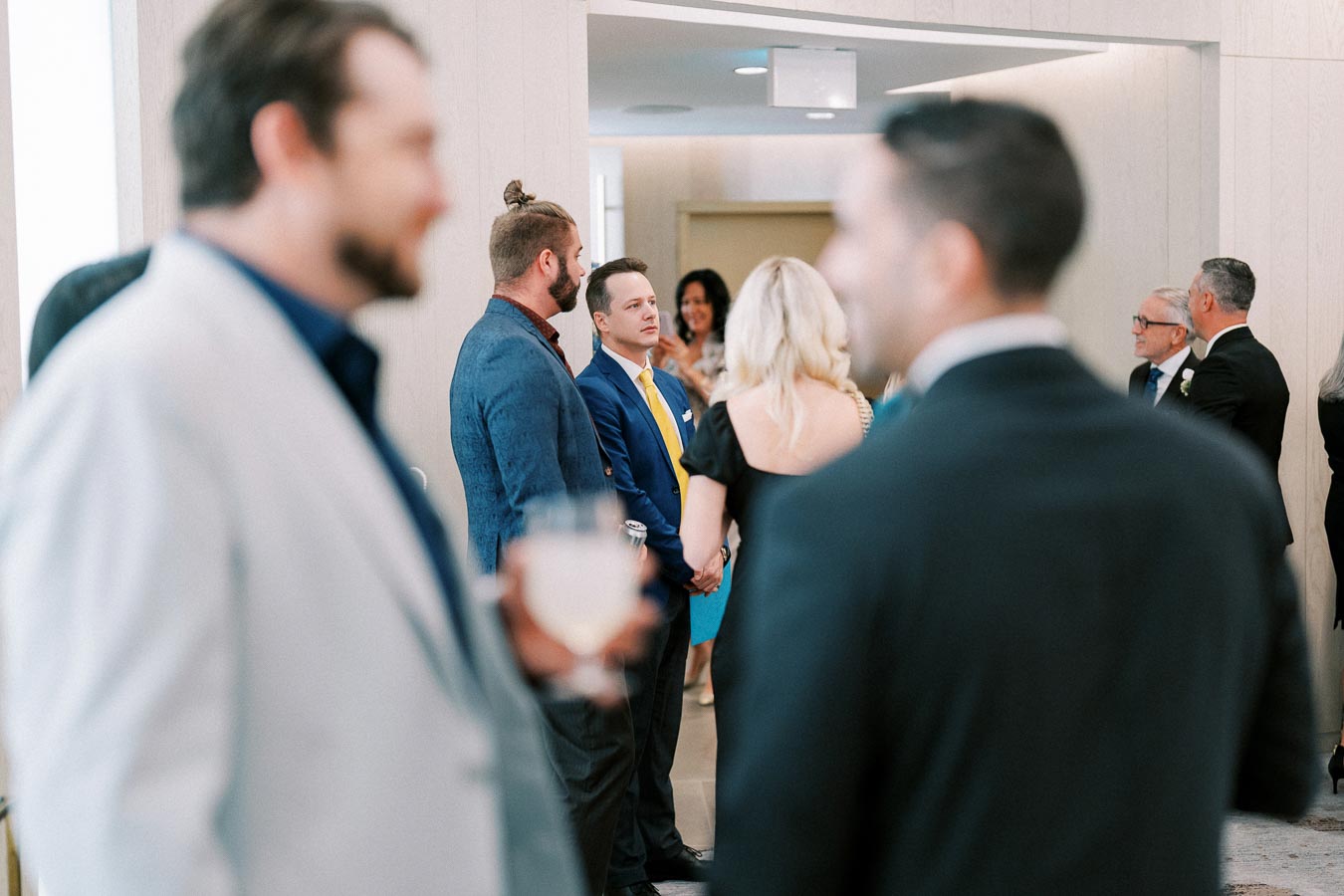 A group of people dressed in formal attire socializing at a sophisticated indoor event, with a man in a blue suit and yellow tie standing prominently in the center.