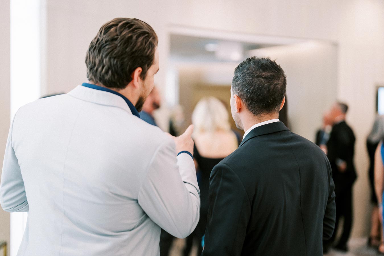 Two men in formal attire engage in conversation at a social event, surrounded by a blurred background of people in formal dresses and suits, suggesting a gathering or networking event.