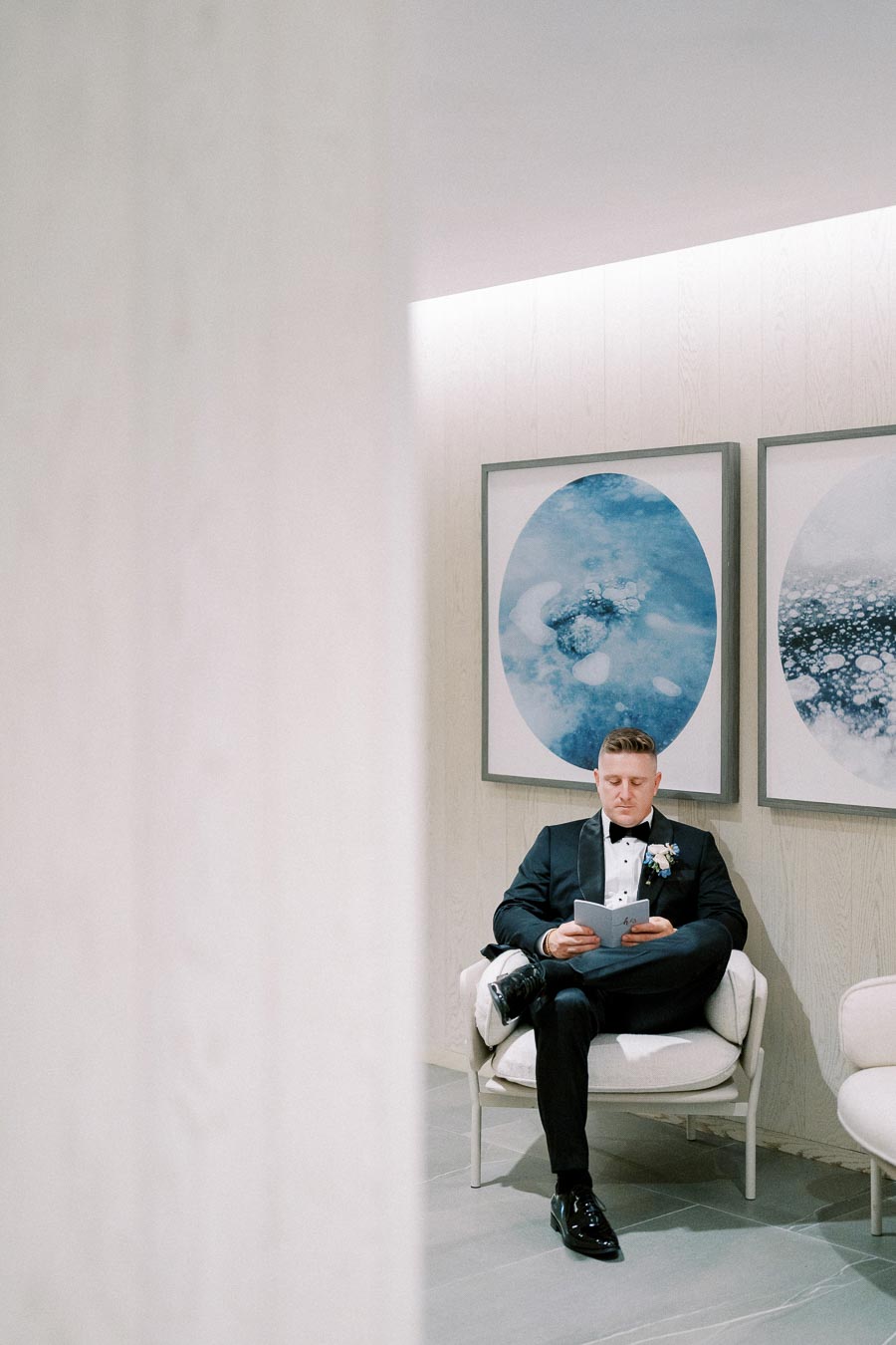 A man in a formal black tuxedo sits in a modern, minimalist interior, reading a booklet. The room features large blue and white abstract art pieces on the wall.