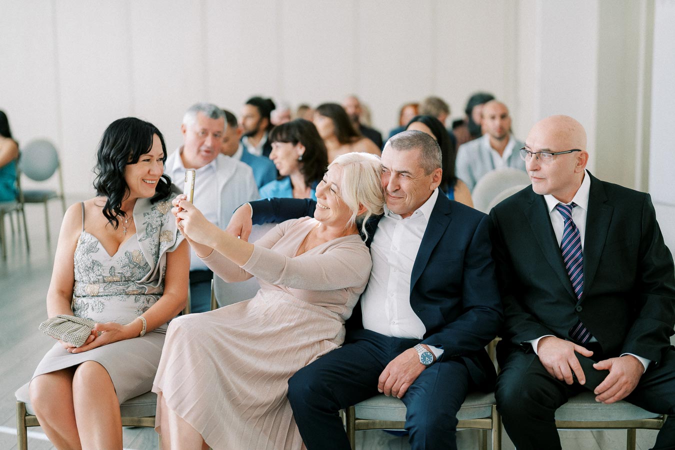 A group of people dressed formally sitting on chairs, with one woman taking a selfie using a smartphone. The setting appears to be a social gathering or event.