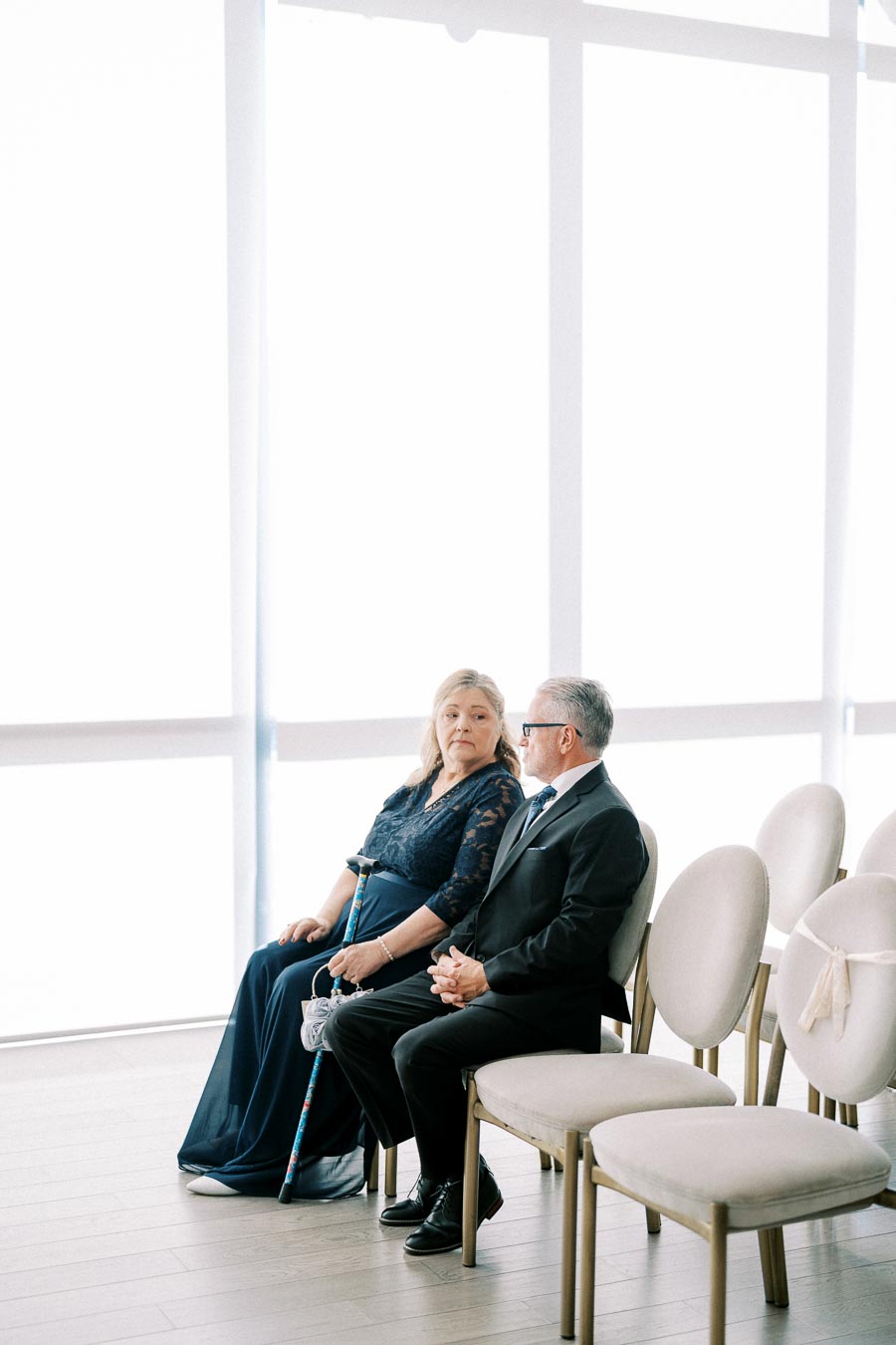 Elderly couple in formal attire sitting on chairs in a brightly lit room, with the woman holding a cane, conveying an elegant and serene atmosphere.