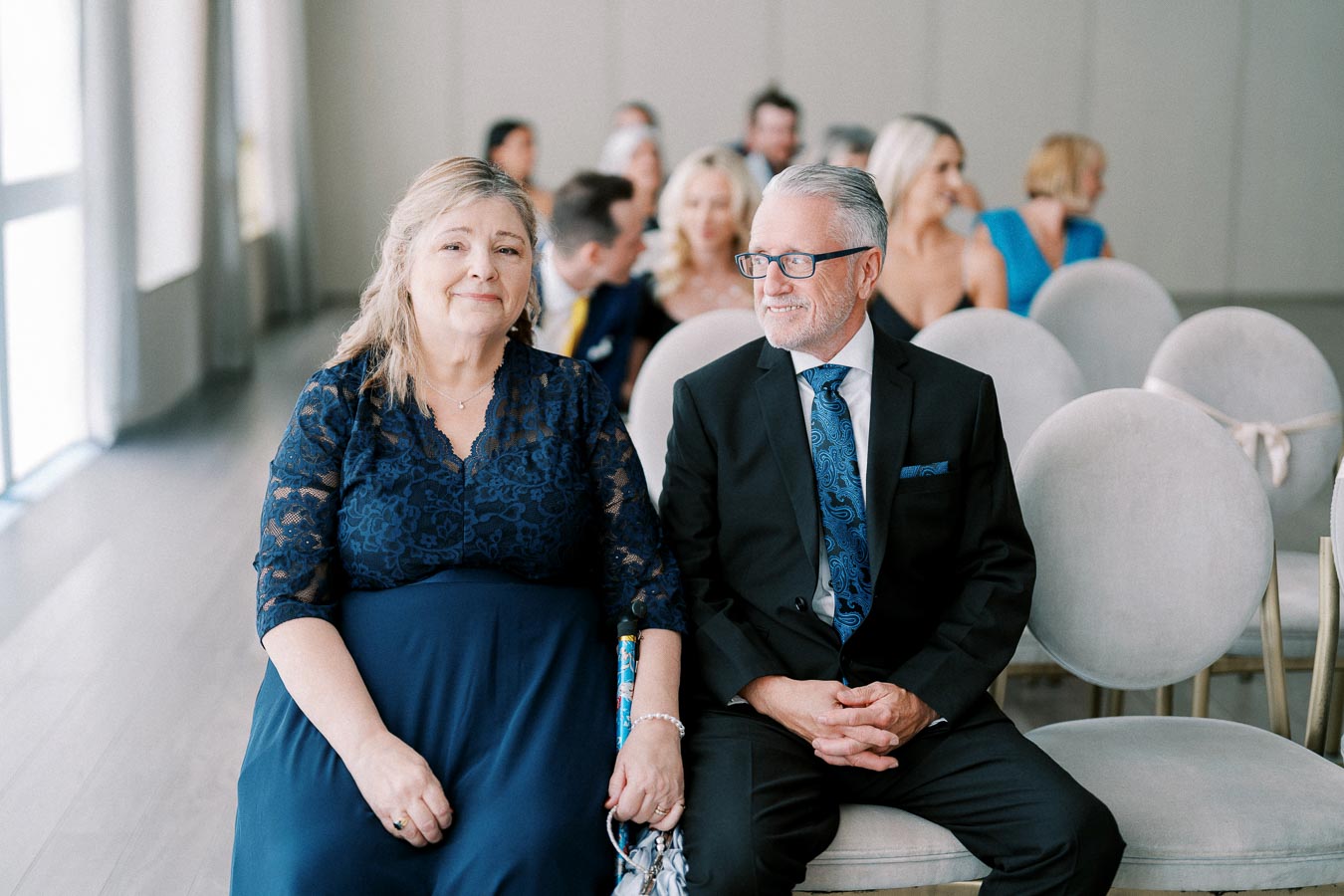 An older couple wearing formal attire seated at an indoor wedding ceremony, with other guests visible in the background.