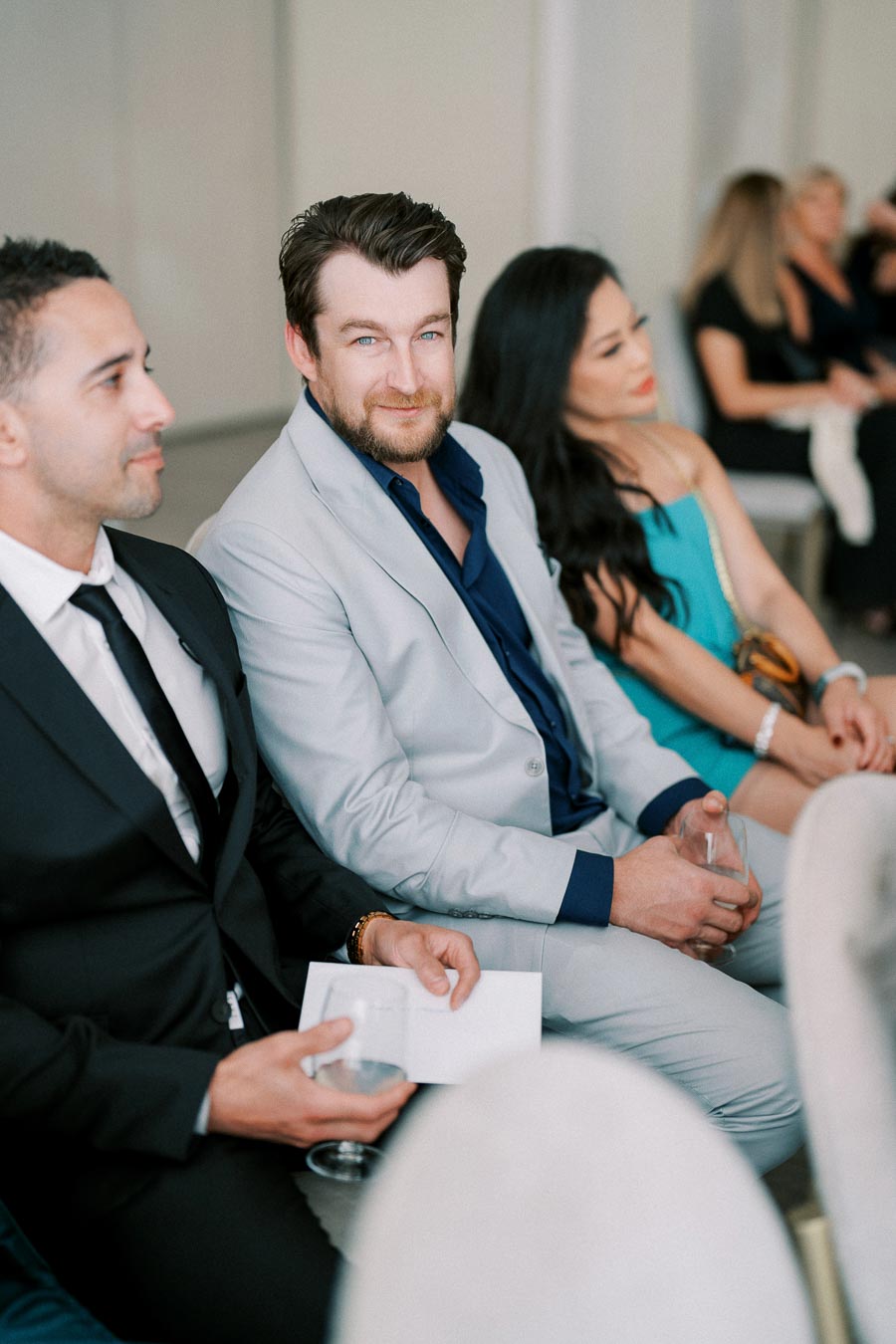 A group of well-dressed individuals seated in a row at a formal event, featuring a man in a light gray suit smiling at the camera, surrounded by two others in stylish attire, creating an atmosphere of elegance and sophistication.