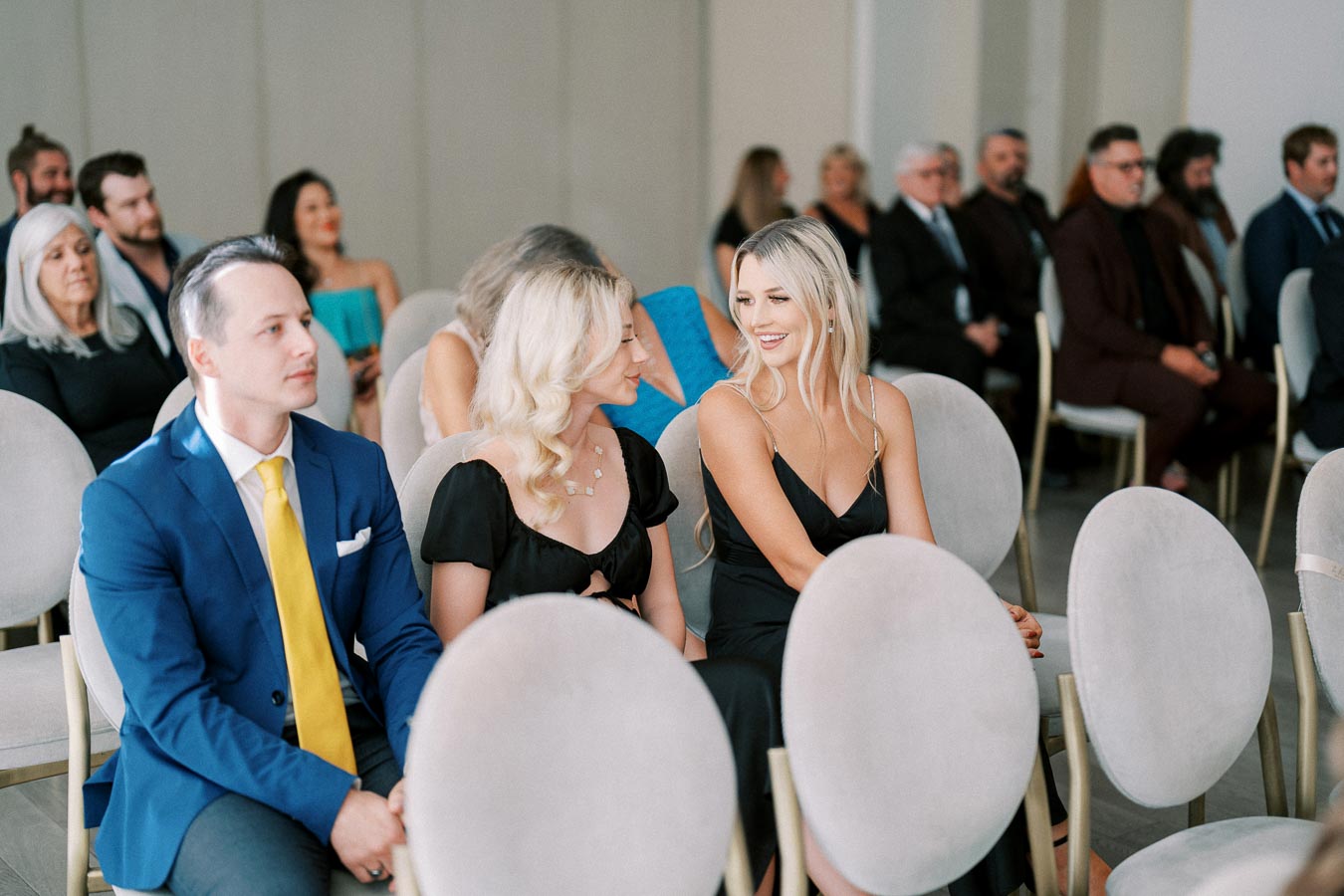 A group of people seated attentively at a formal event, with two women in elegant black dresses conversing and a man in a blue suit with a yellow tie sitting beside them, surrounded by other guests in a well-lit venue.