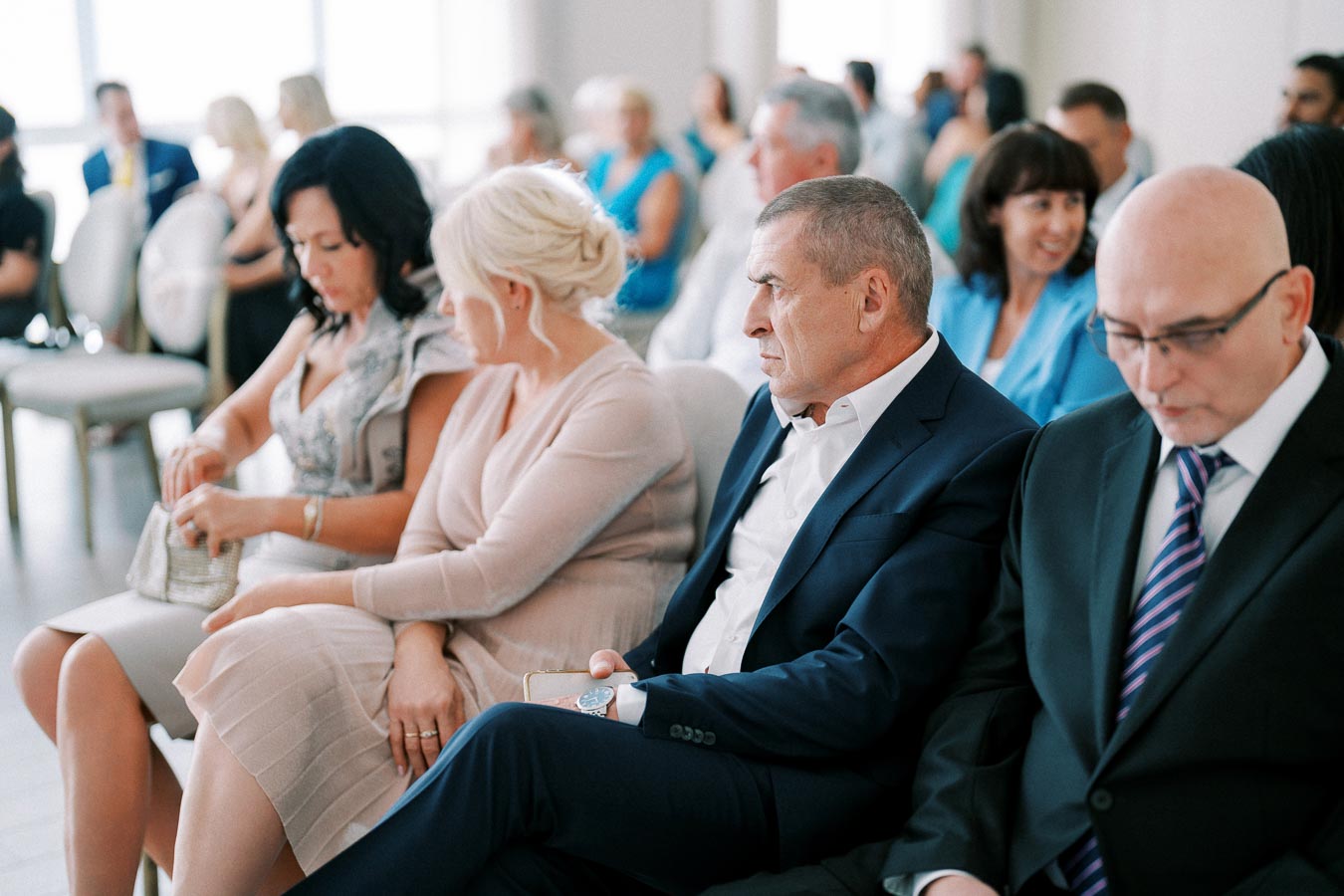 Group of people seated at a formal event, focusing on two men in suits alongside women in elegant attire, engaged in conversation.