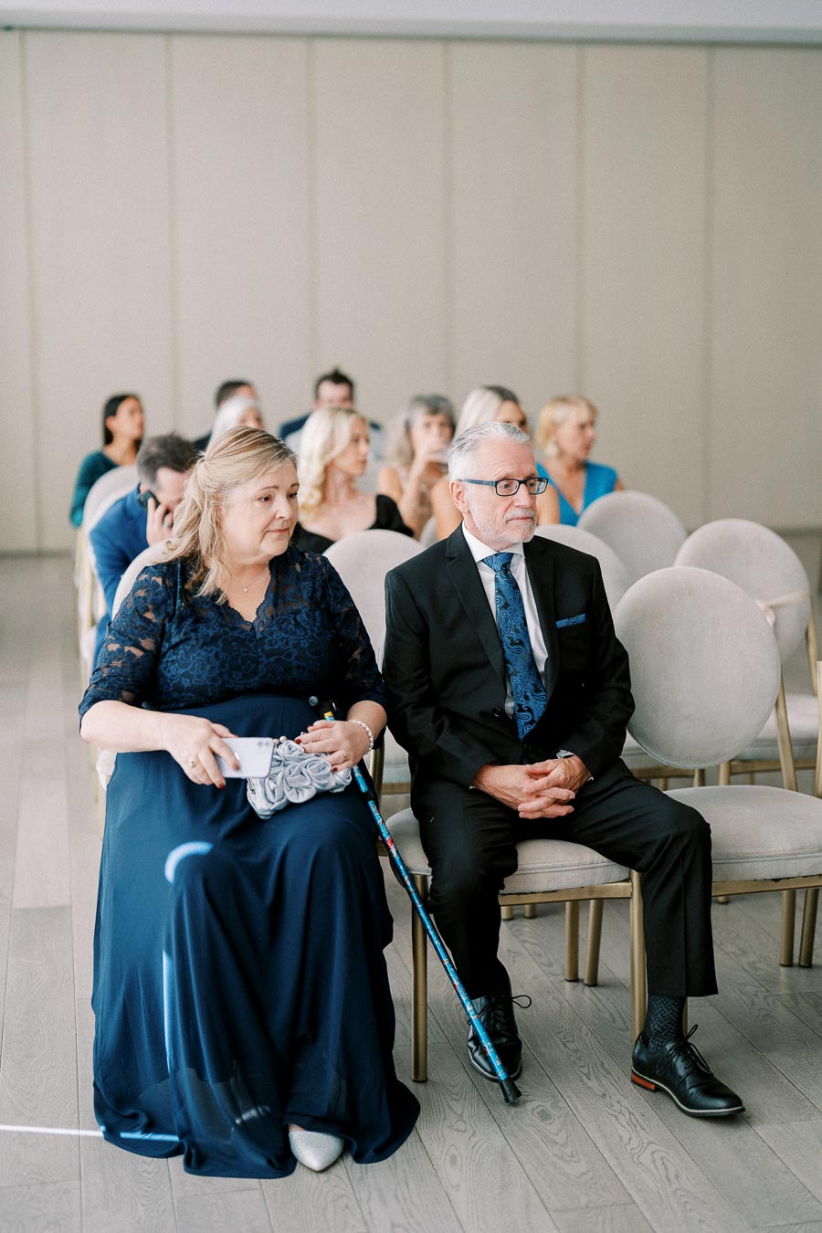 Elderly couple dressed in formal attire sitting in a row of chairs at an indoor event, surrounded by other attendees.